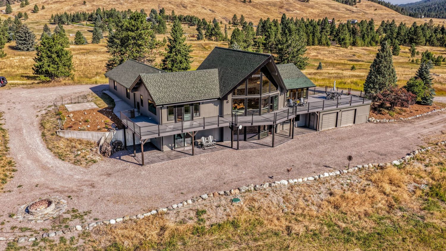 Aerial view of a large mountain home with a wraparound deck, green roof, and scenic rolling hills with pine trees.
