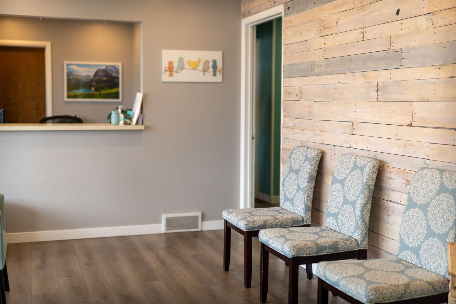 Modern waiting room with light wood accent wall, two patterned chairs, and a reception desk in a cozy office.