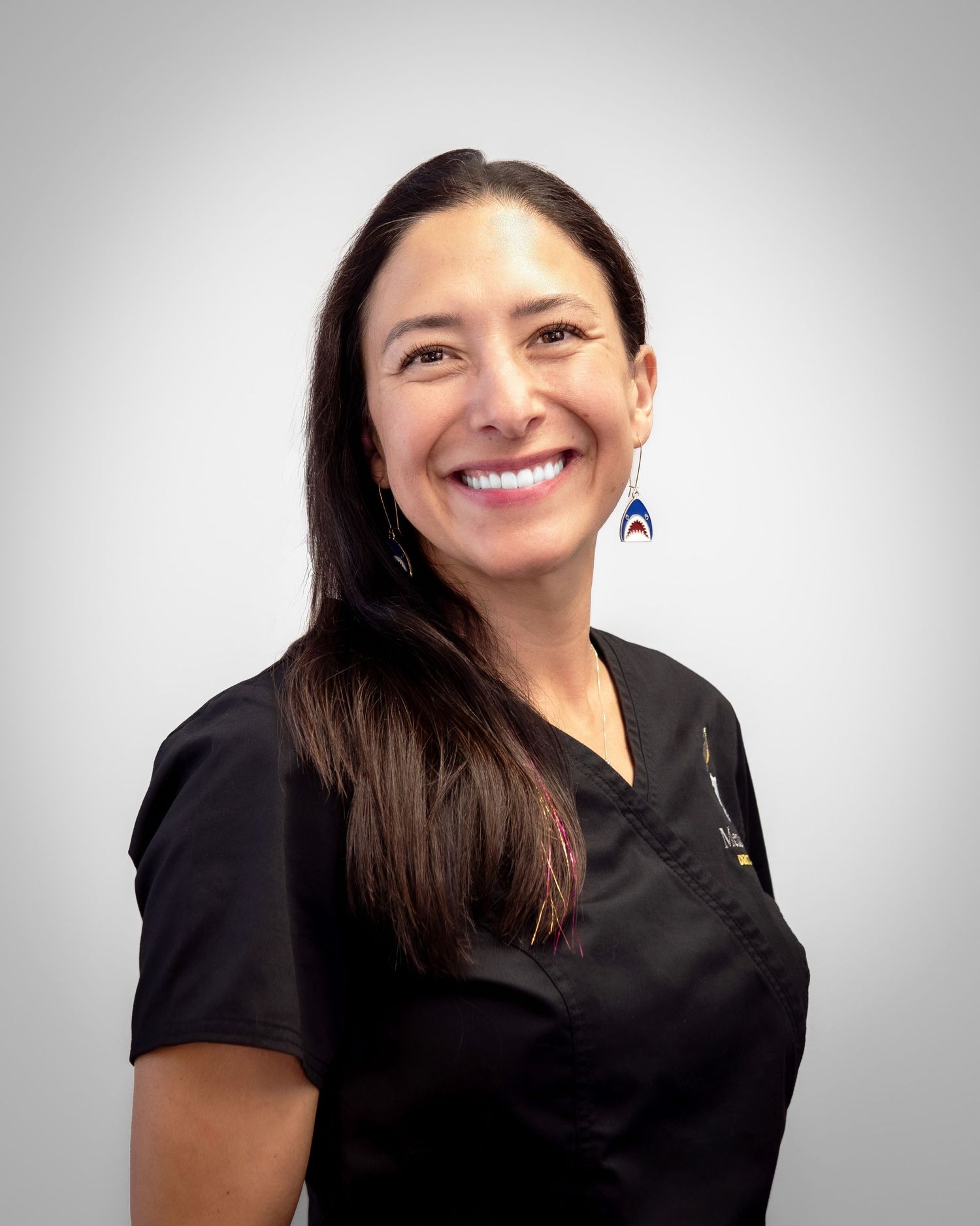 Smiling female healthcare professional in black scrubs with long dark hair and blue earrings against gray background.
