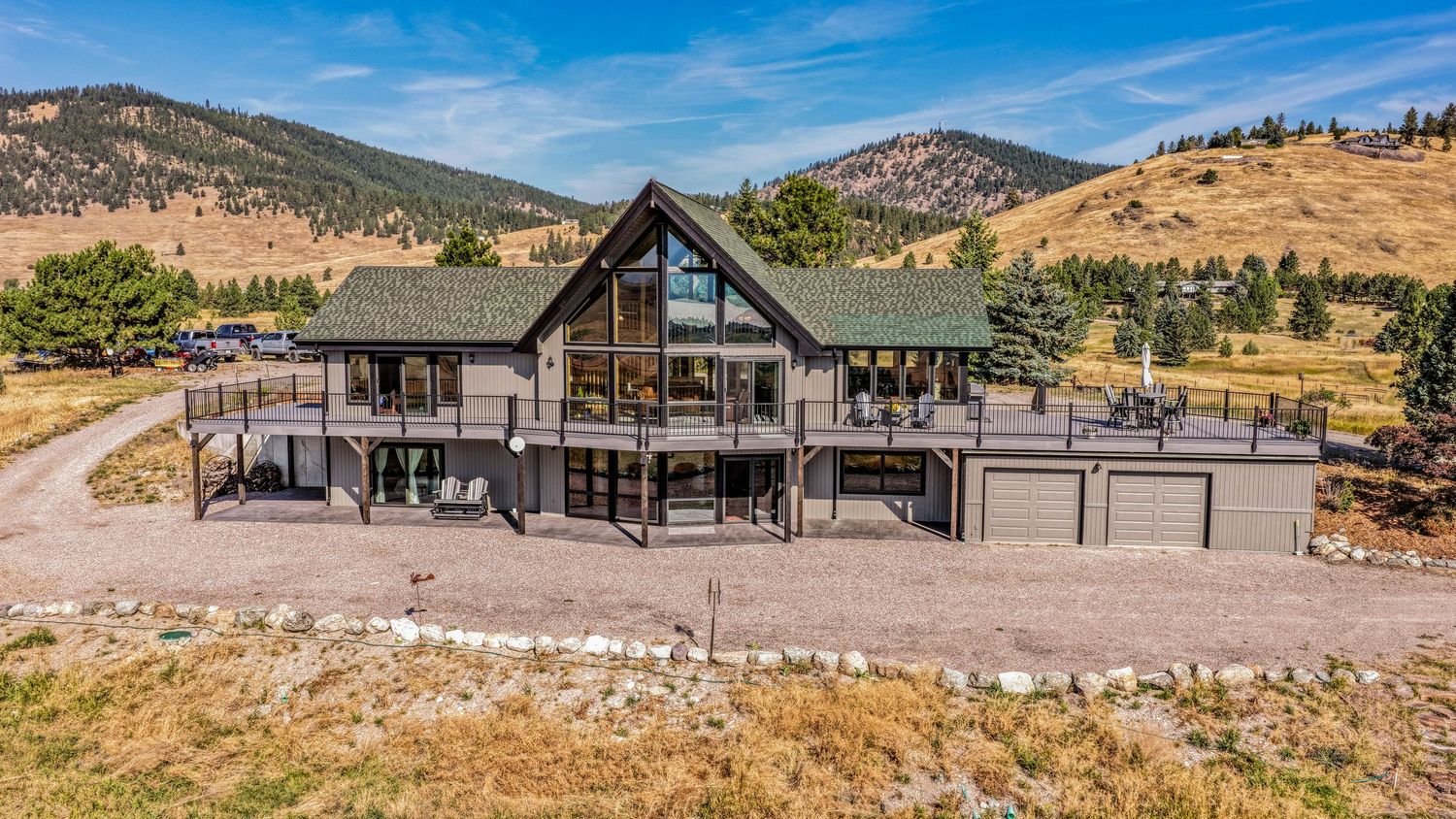 Large mountain home with green roof, expansive deck, and three-car garage set against rolling hills.