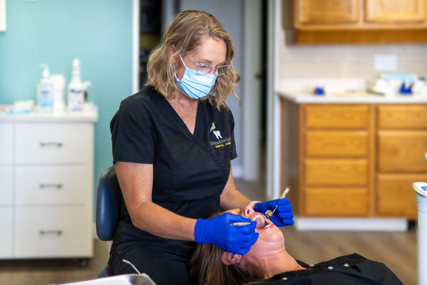 Female dental hygienist in black scrubs and blue gloves performing a dental procedure on a patient.