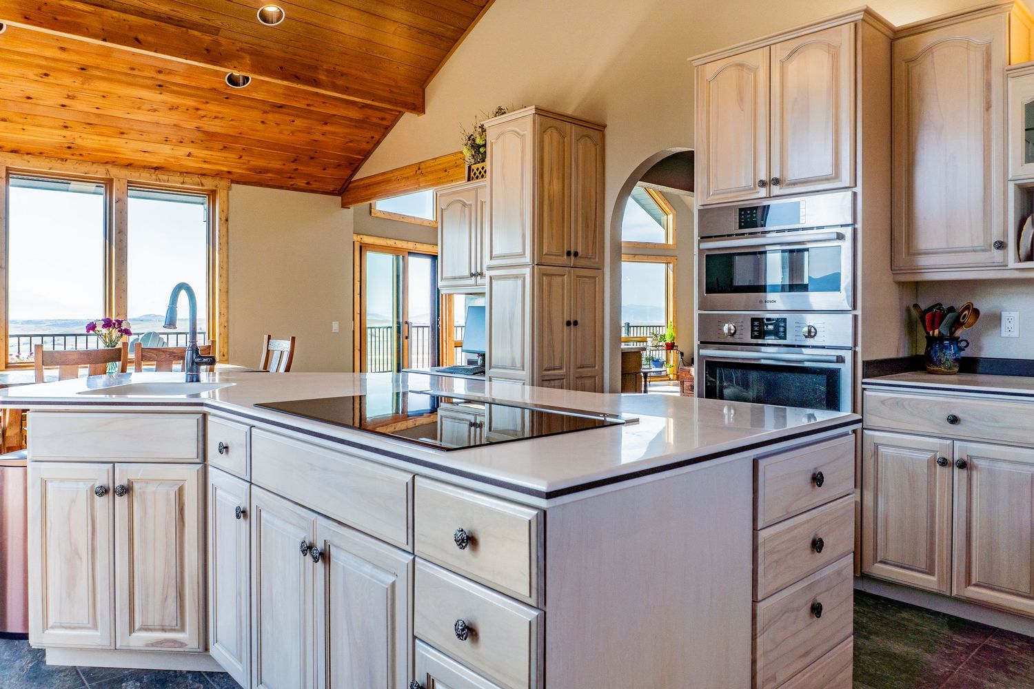 Spacious kitchen with white cabinetry, black cooktop island, stainless steel ovens, and vaulted wood ceiling.