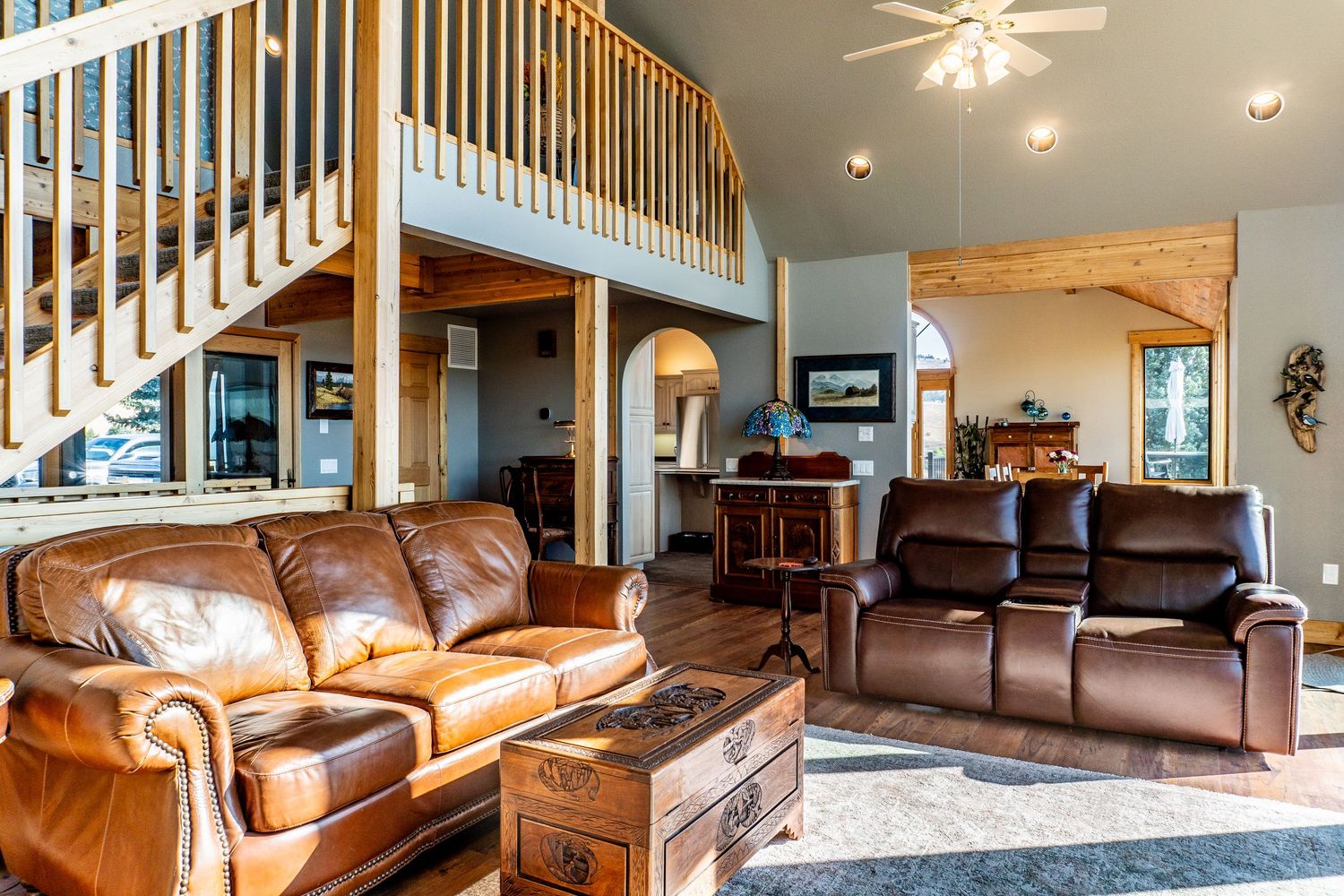 Spacious living room with leather sofas, wood staircase, loft, and high vaulted ceilings in a rustic home.