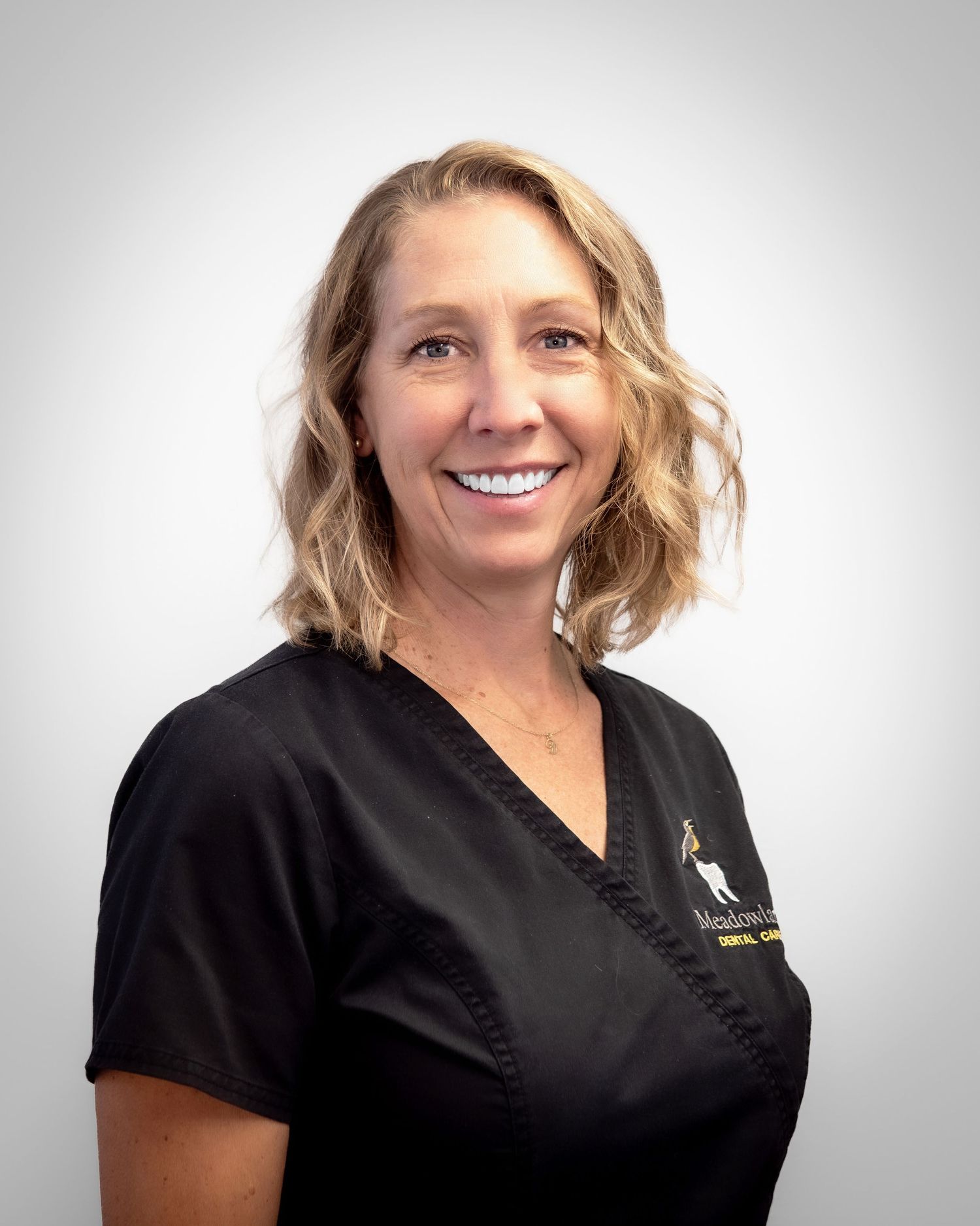 Smiling female dental professional in black scrubs with logo, blonde wavy hair, professional headshot photo.