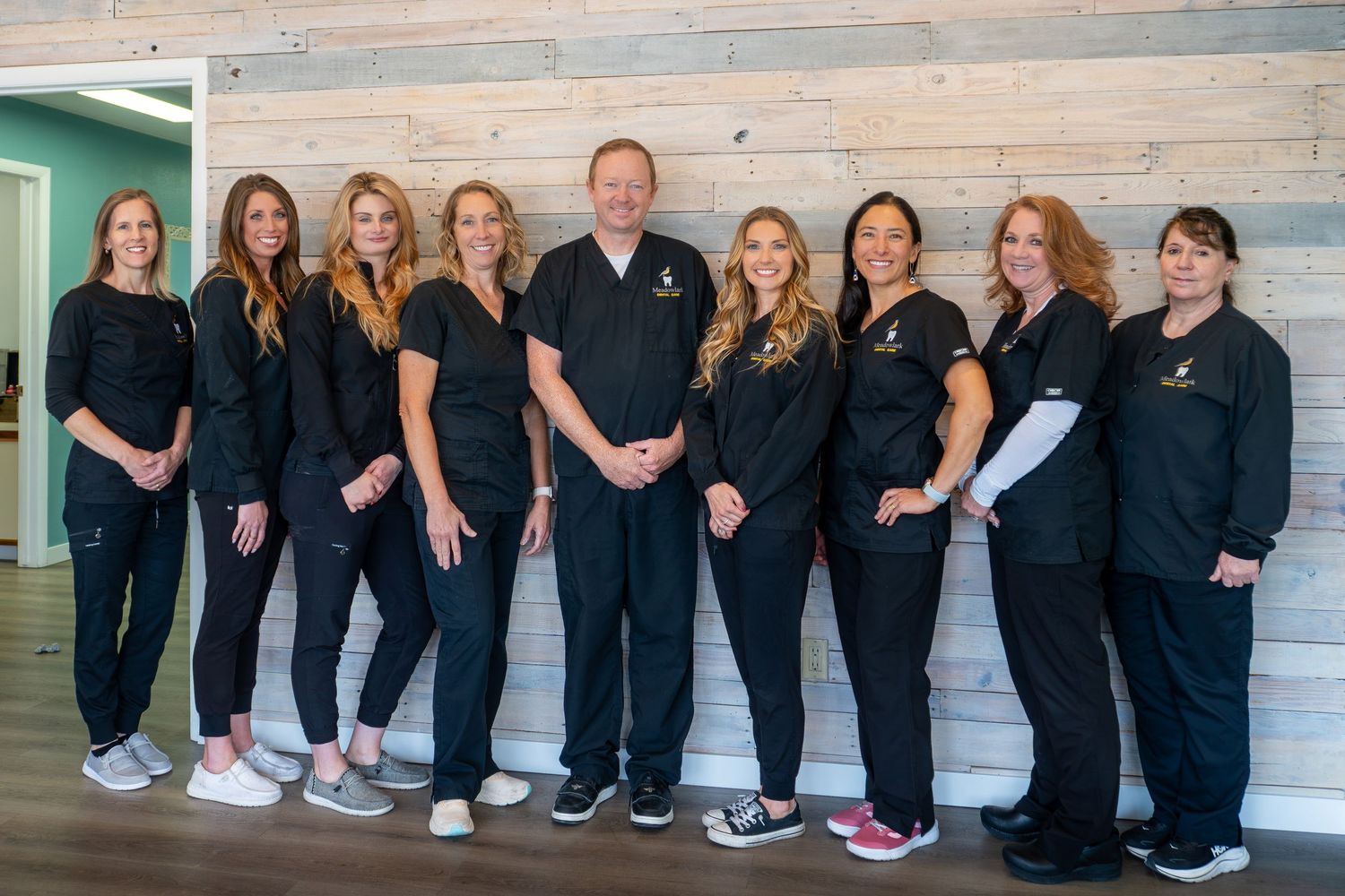 Dental team in black scrubs posing together in a modern clinic with wood panel walls.