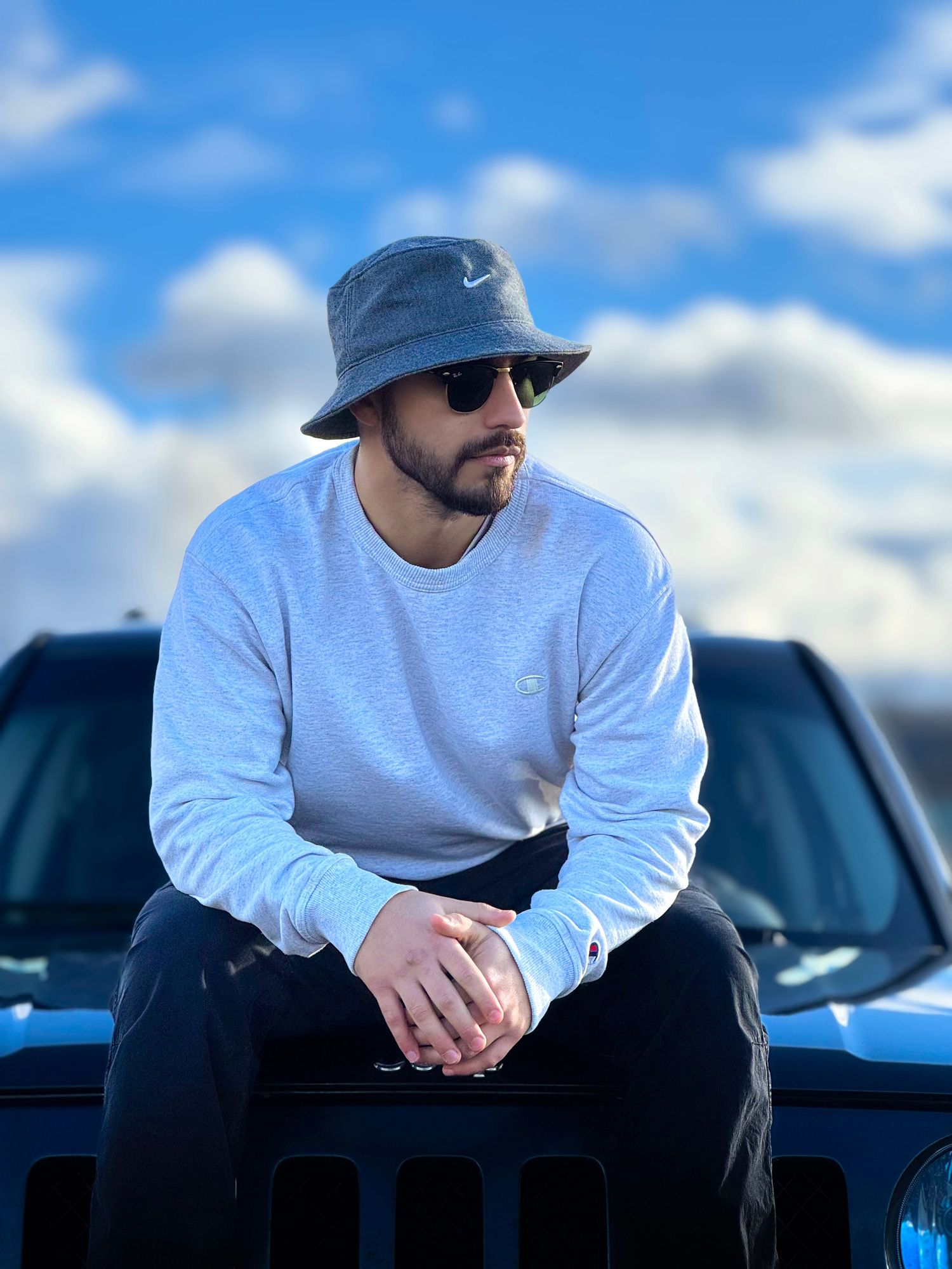 Man in gray sweatshirt and bucket hat sits on blue Jeep hood under cloudy sky.