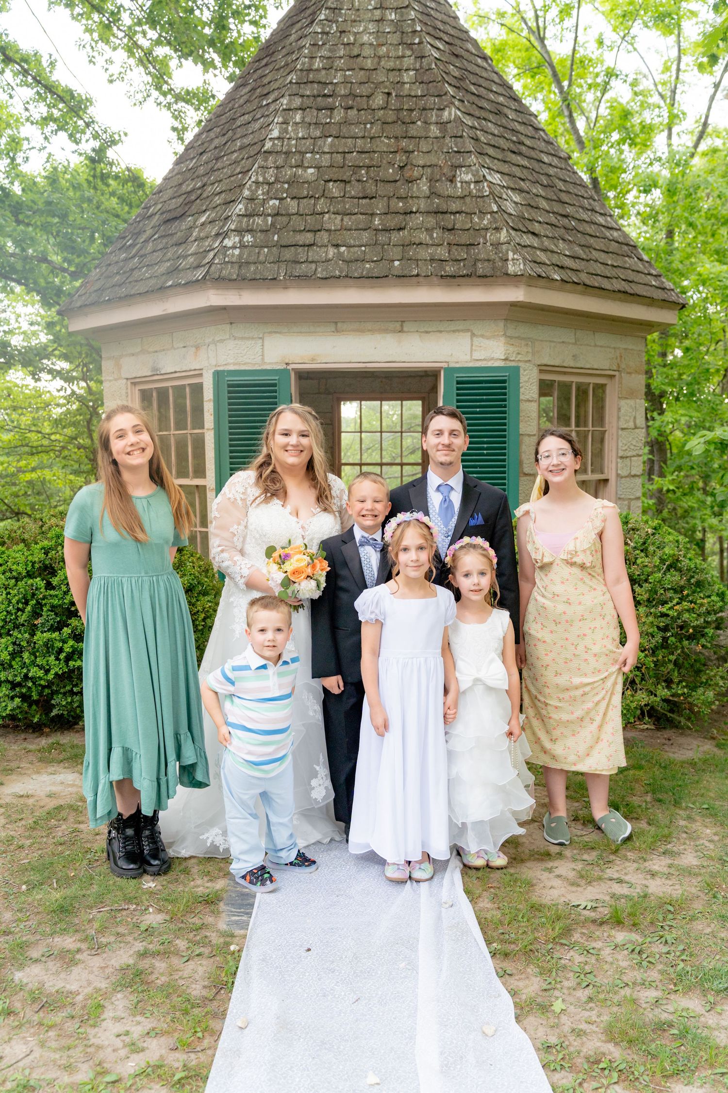 Wedding party poses before a charming gazebo, bride holding bouquet with groom and family including children in formal attire. Row and Co