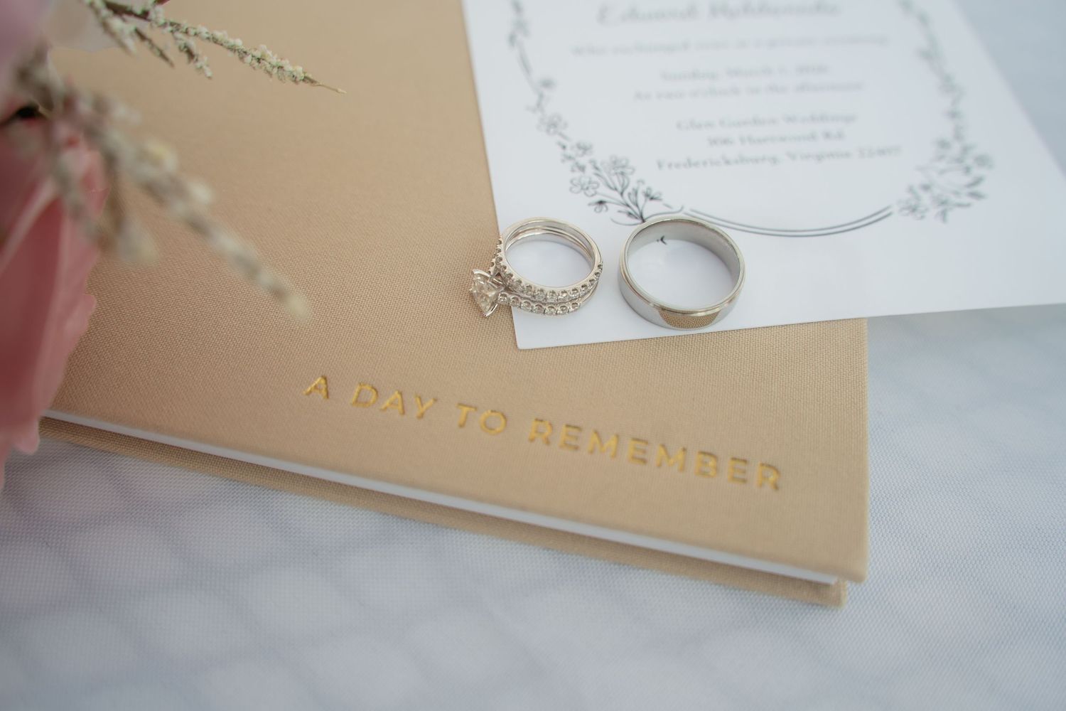 Wedding rings resting on a gold embossed journal reading A Day To Remember beside an invitation and pink flowers.