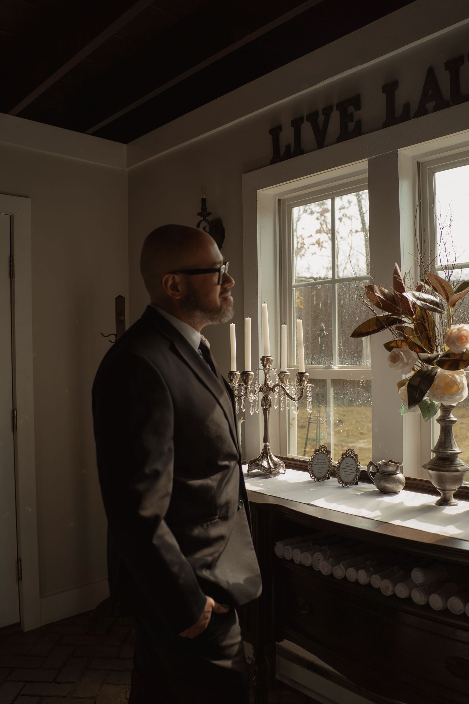 Bald man in dark suit with glasses gazing out window beside candelabra and floral arrangement in elegant room.