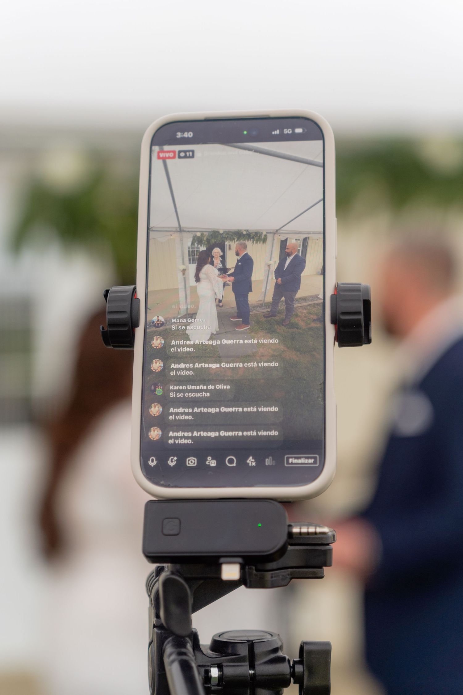 Smartphone on tripod live streaming a wedding ceremony outdoors under a tent, with blurred guests in the background.