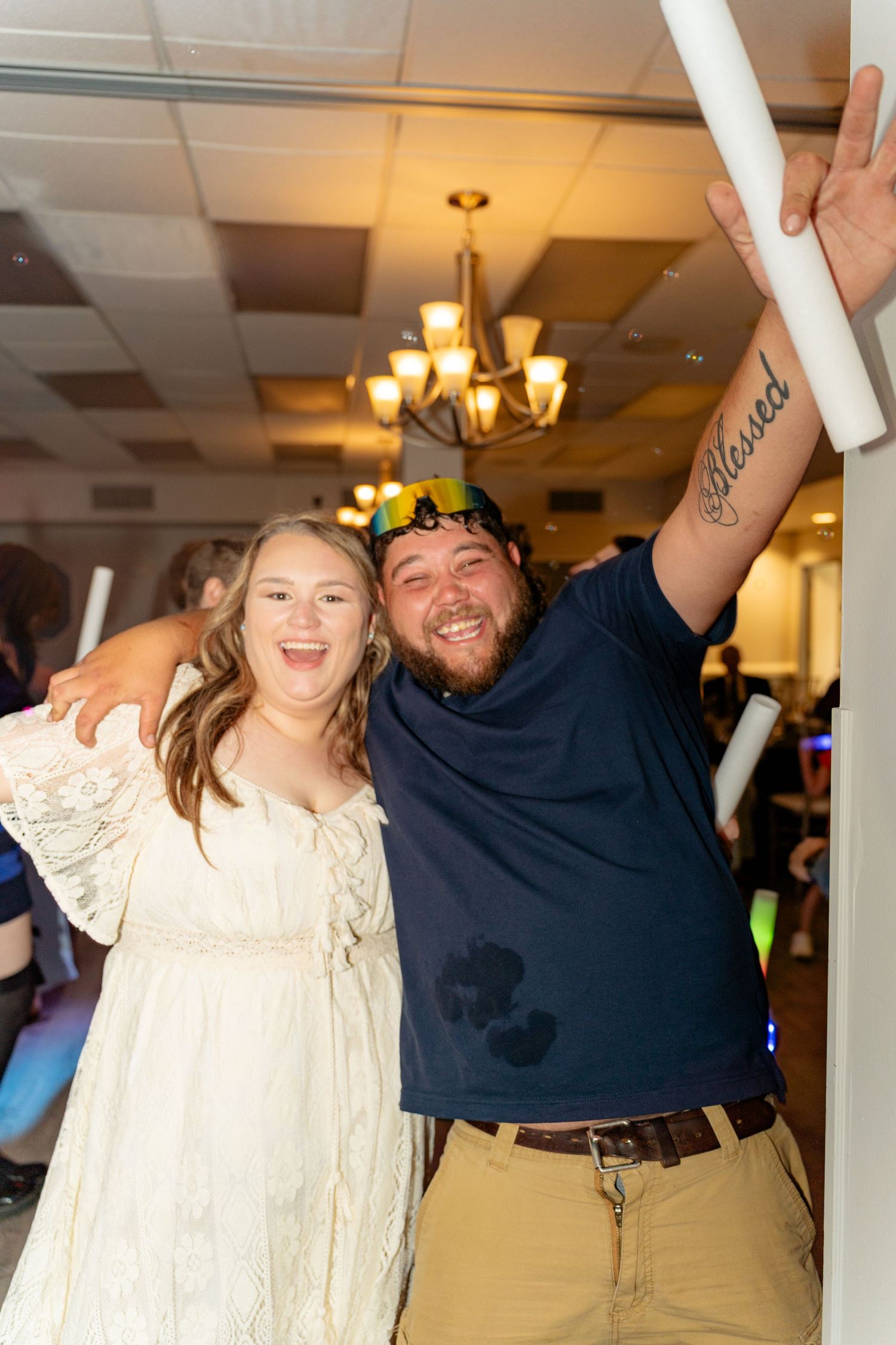 Two friends smile and pose together at a wedding reception, man raising arm showing Blessed tattoo.