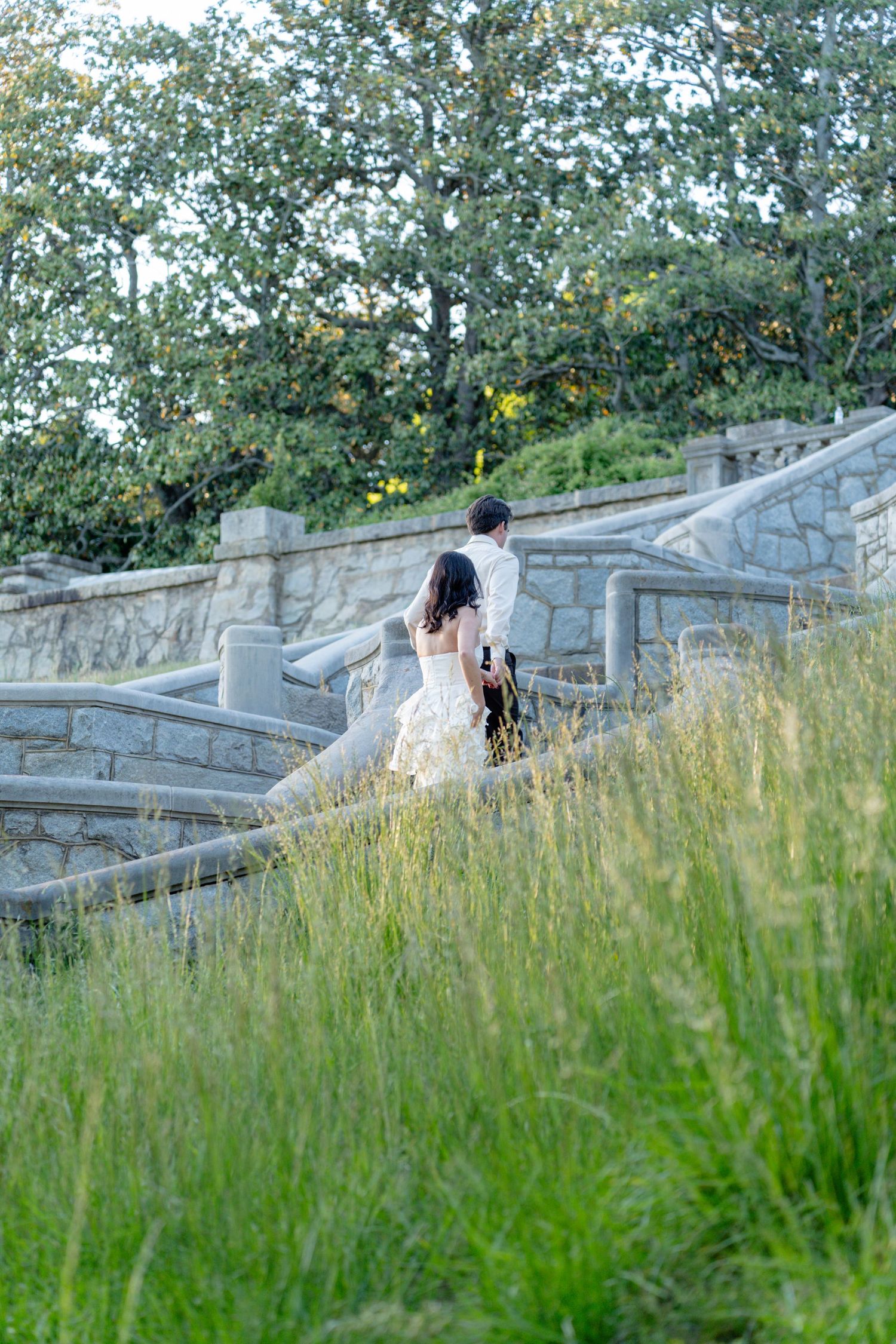 Bride and groom walking up stone stairs through tall golden grass and lush green trees at sunset. Taken by Row And Co Photography in FXBG VA
