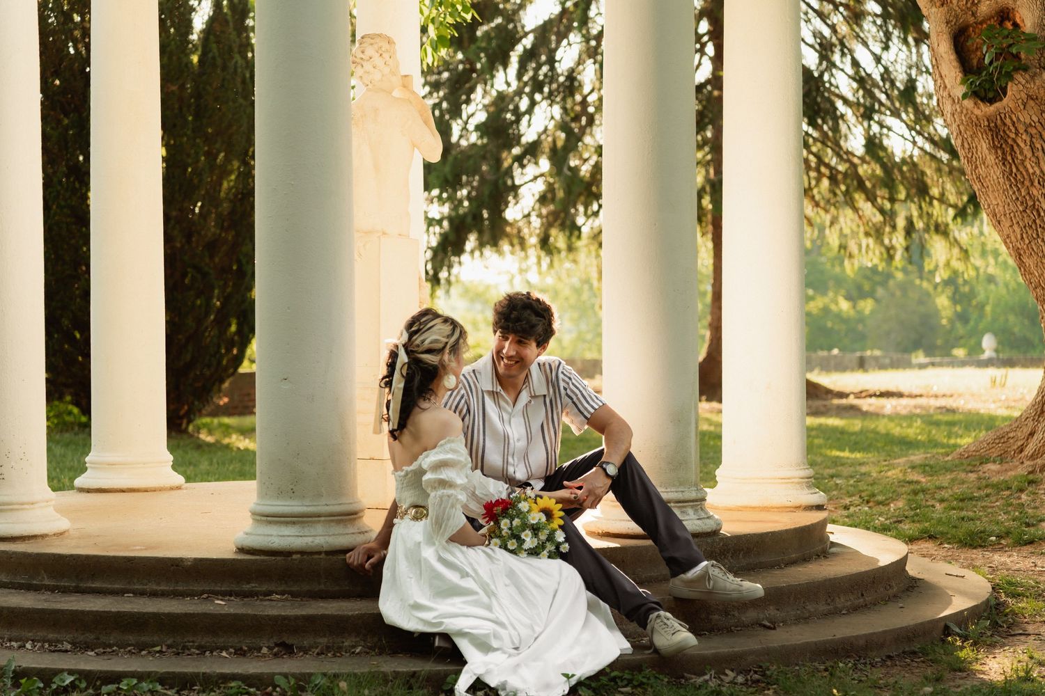 A bride and groom sit on white columned steps surrounded by lush greenery, sharing a romantic moment with a colorful bouquet. Taken in FXBG
