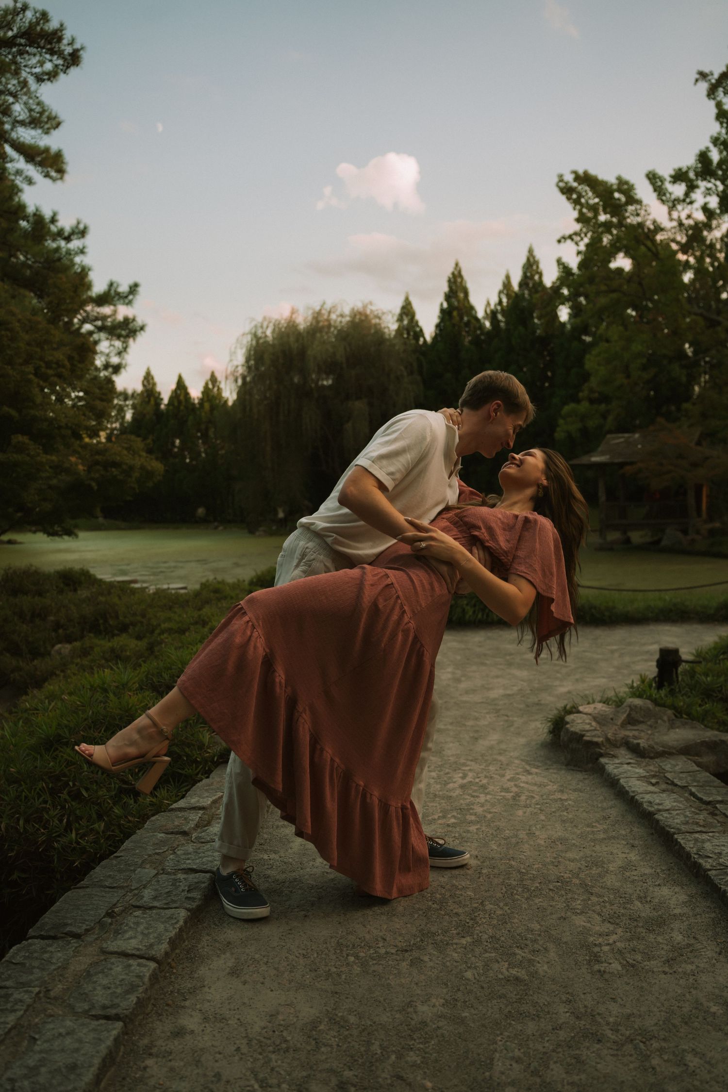 Couple sharing a romantic dip kiss on a stone path in a lush green park at dusk, woman in rust dress.
