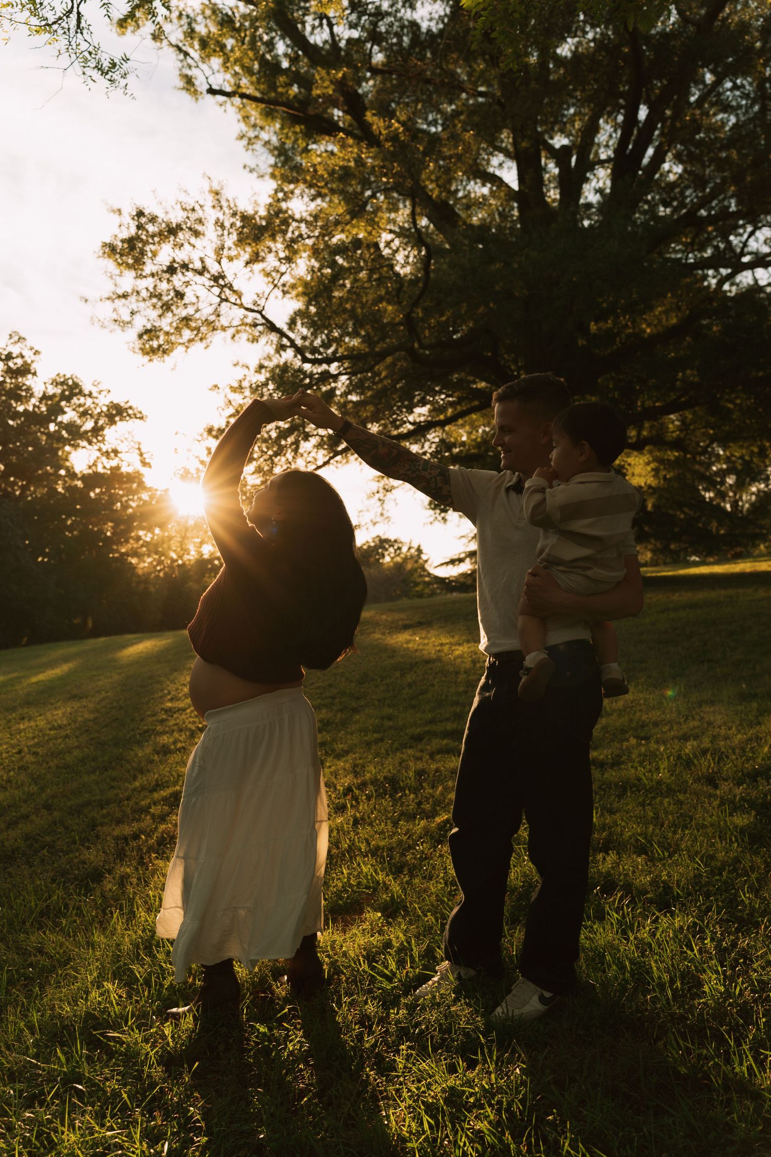 A family dances together in a sunlit park at golden hour, with parents holding their child outdoors.
