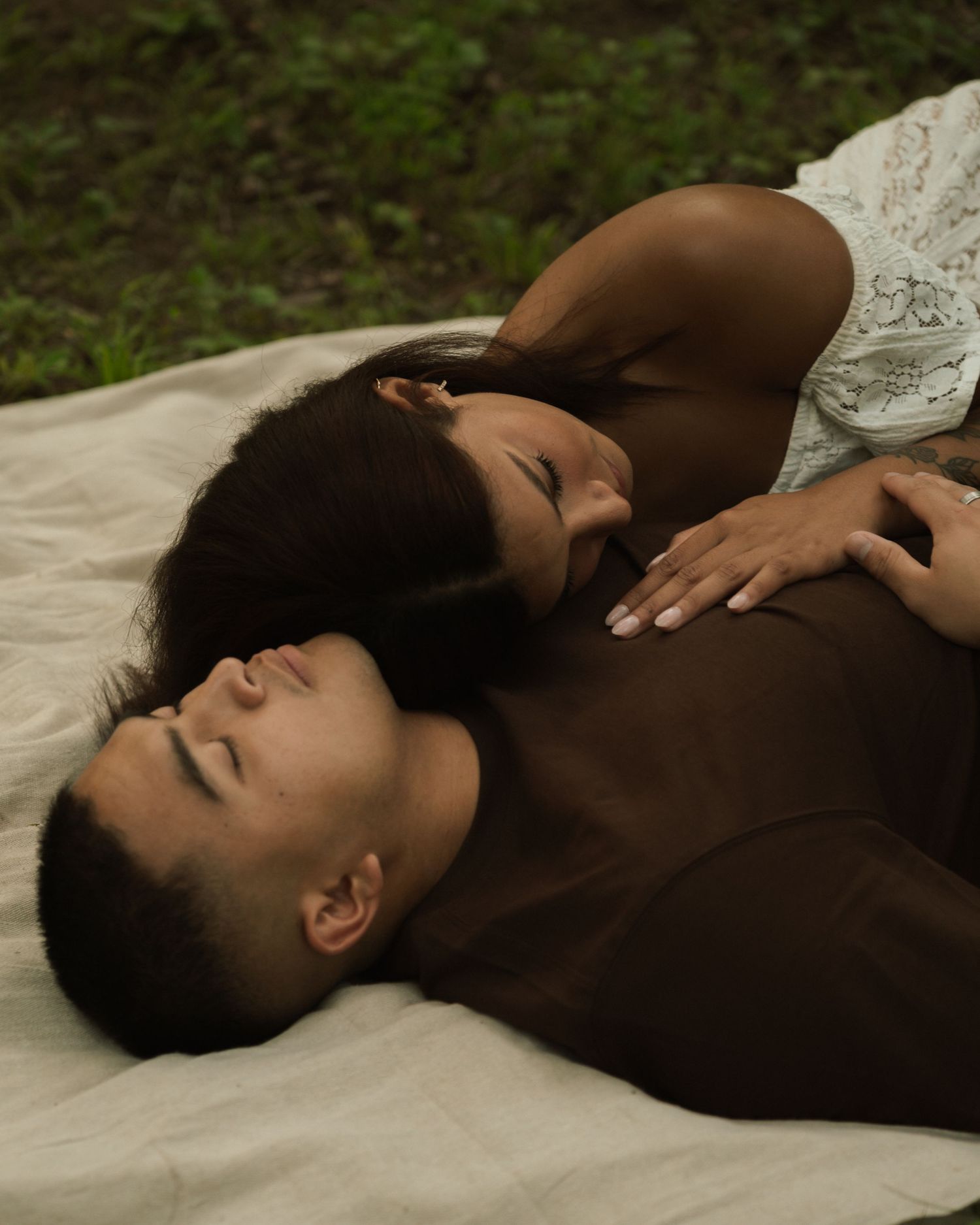 A couple lies peacefully on a blanket outdoors, the woman resting her head on the man's chest in a serene embrace.