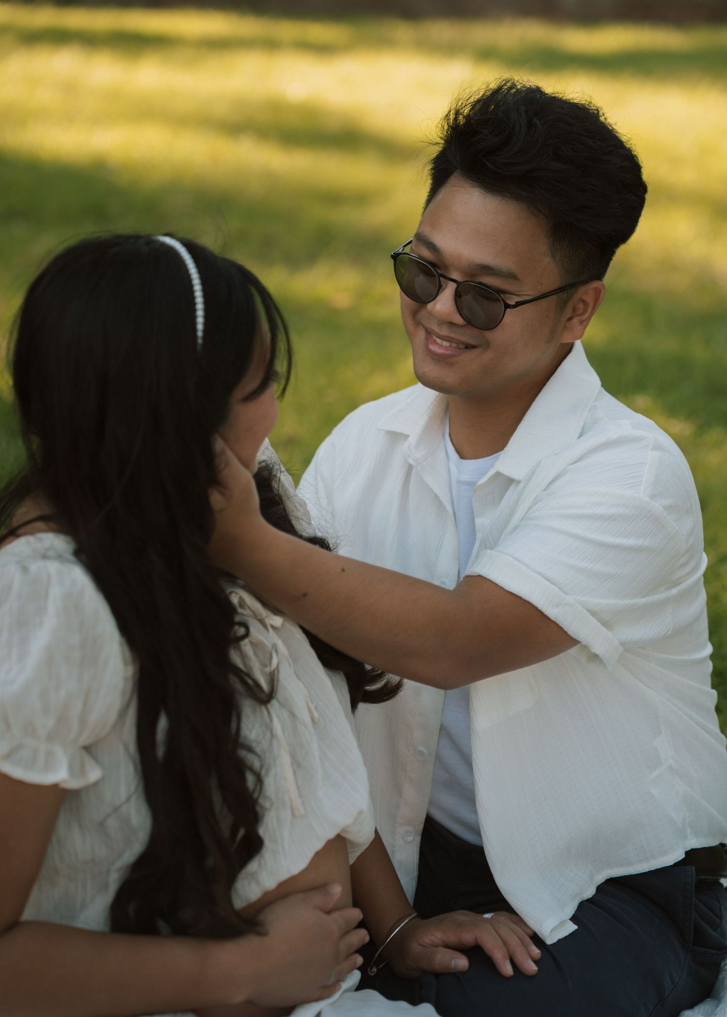 A smiling man in sunglasses and white shirt sits with a woman in a white dress on green grass.