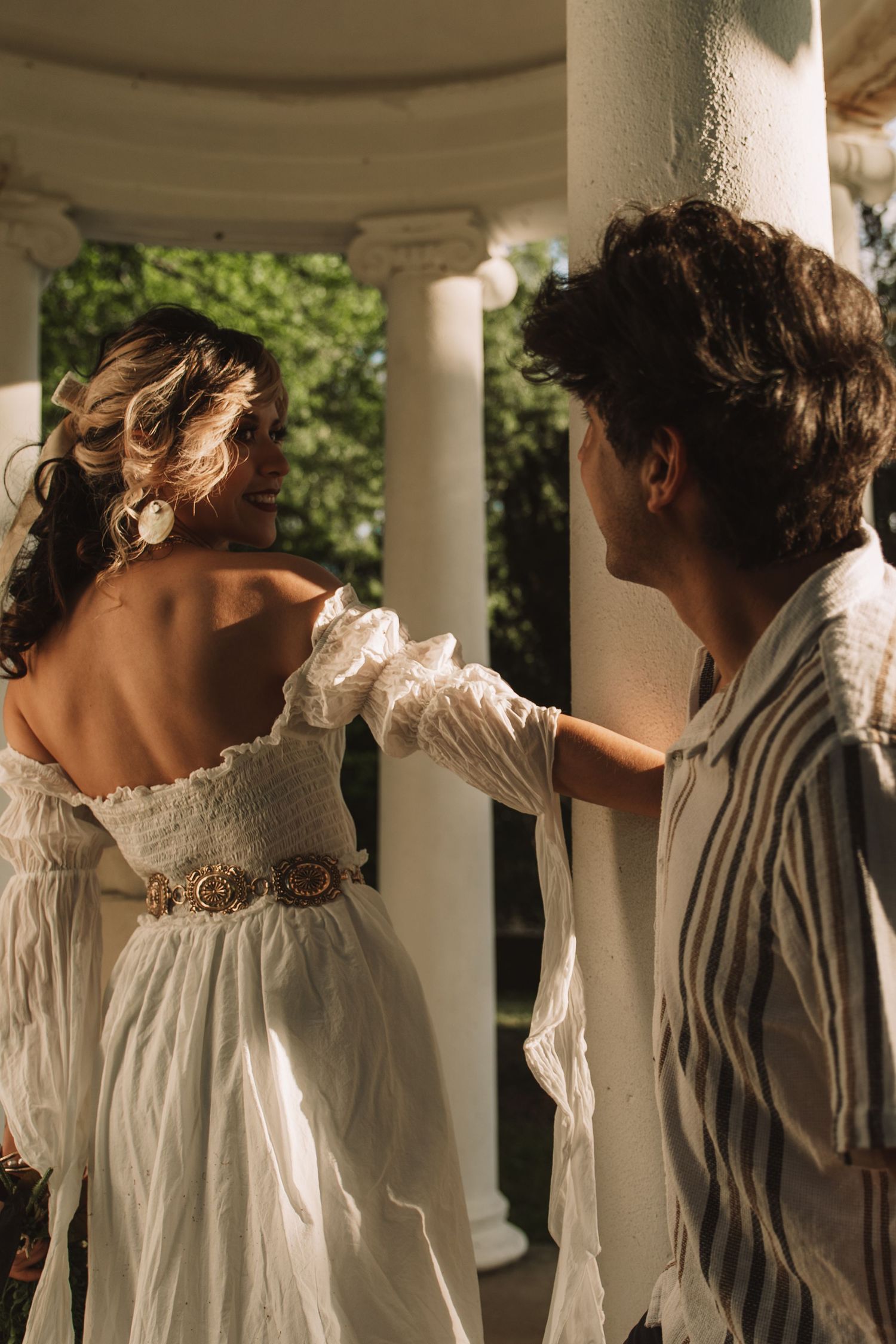 Couple in romantic outdoor setting, woman in white off-shoulder dress with gold belt, man in striped shirt by white columns. Taken by Row&Co
