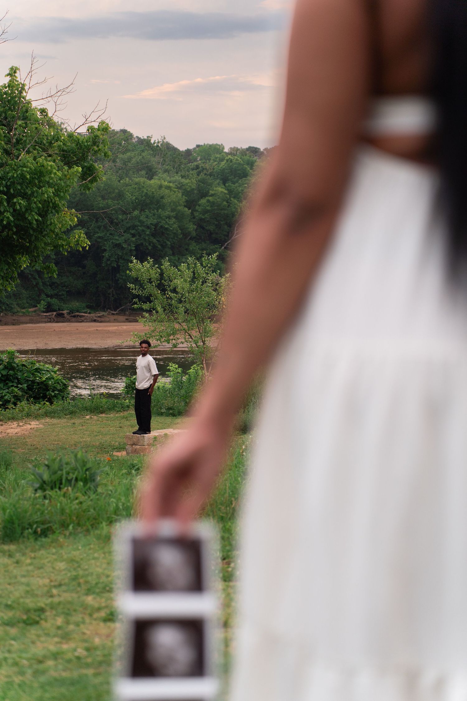 Pregnant woman in white dress holds ultrasound photos while partner stands in background near river surrounded by green trees.