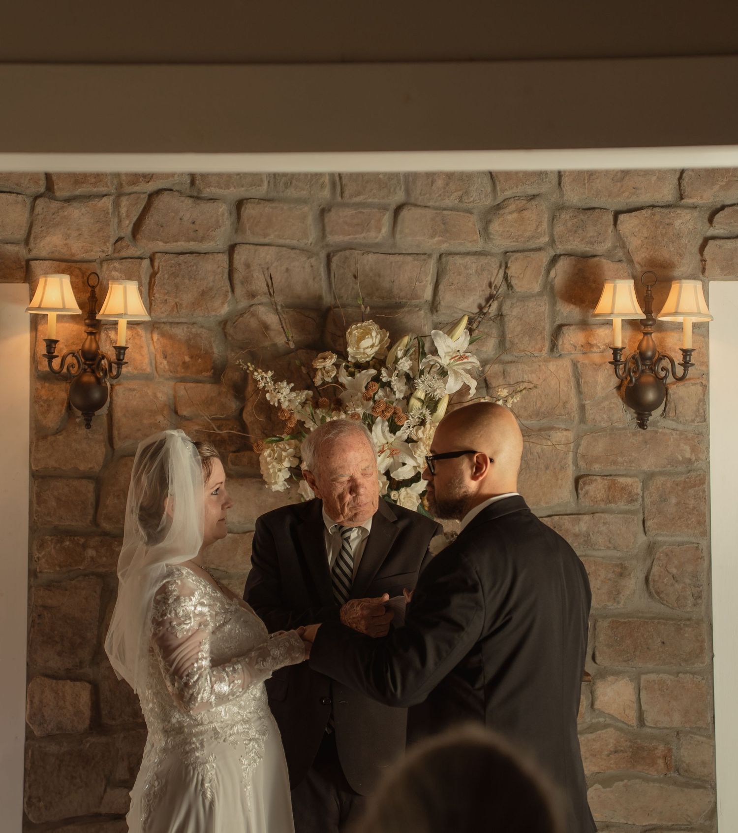 A bride and groom exchange vows before an officiant at a rustic stone wall altar adorned with white flowers and wall sconces.