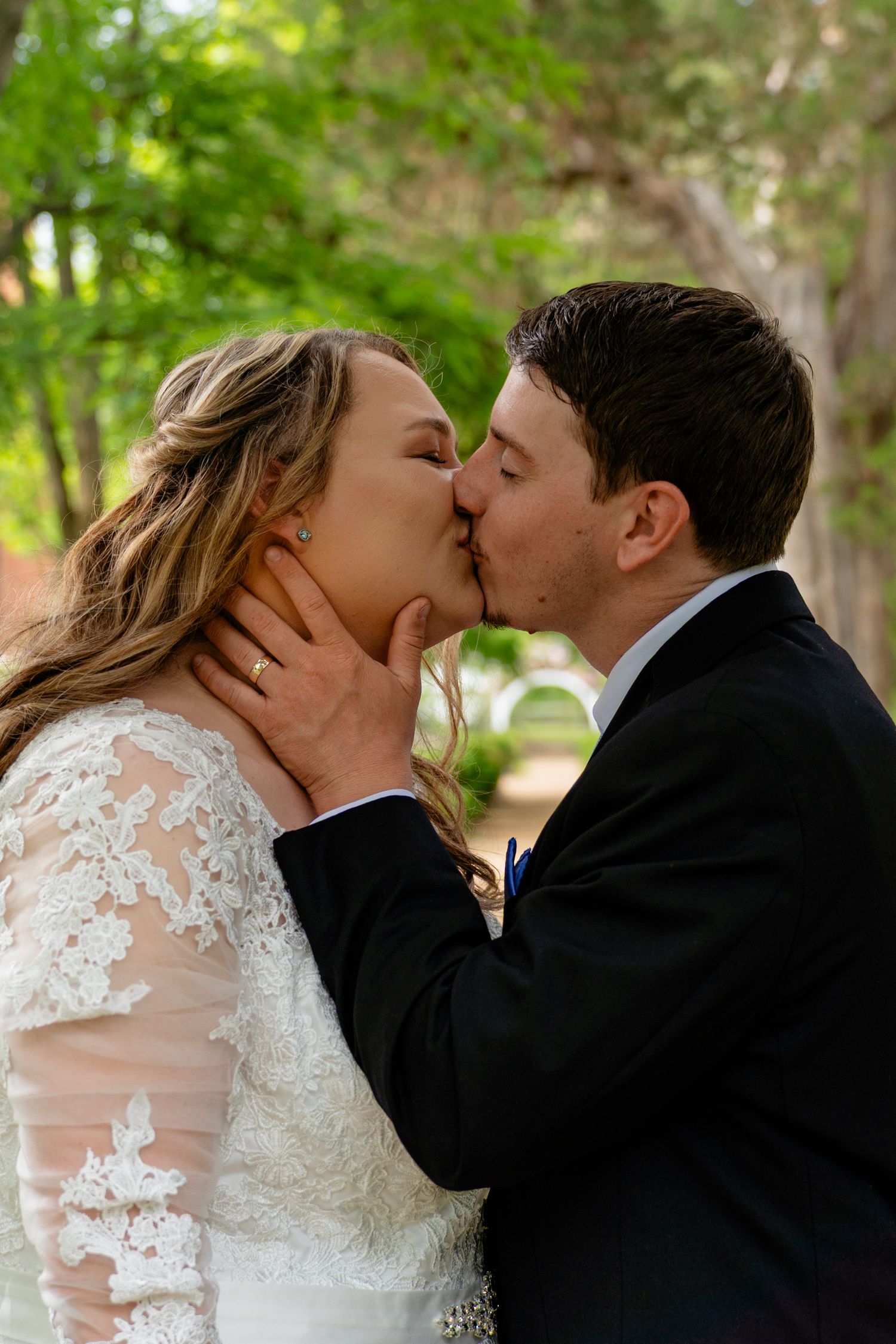 Bride and groom share their first kiss as a married couple outdoors surrounded by lush green trees.