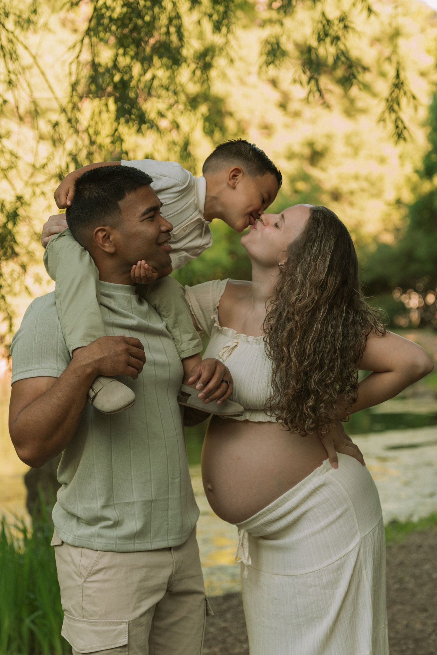 Pregnant woman and man with child on shoulders share a tender family moment outdoors in a lush green park setting.
