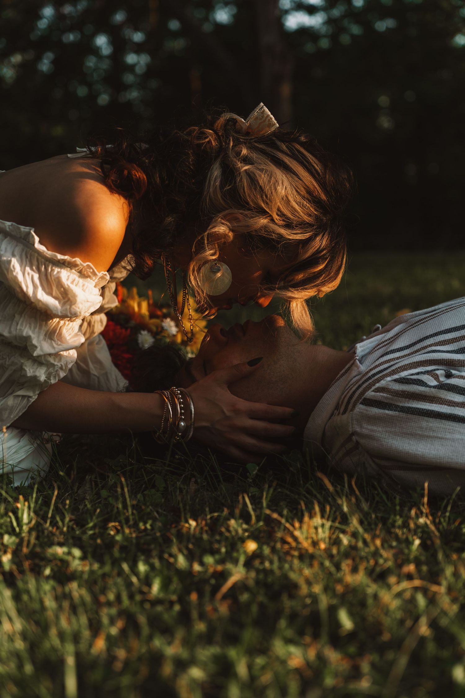 A woman with a floral crown leans over a person lying in golden sunlit grass, holding a bouquet in a romantic outdoor setting.
