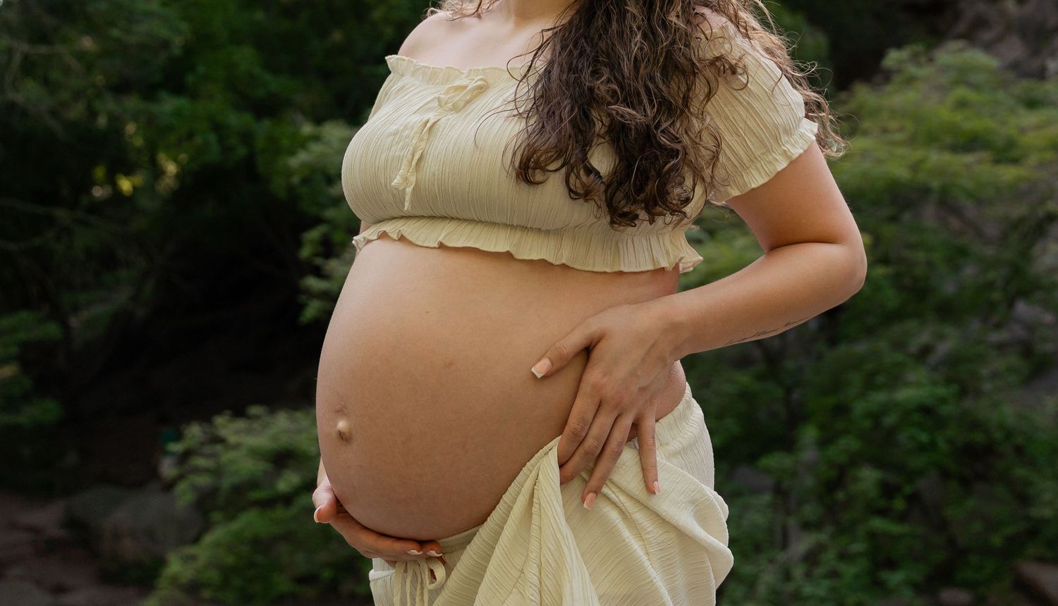 Pregnant woman in yellow dress cradling her bare baby bump outdoors among green foliage. Taken by Row And Co Photography in FXBG VA