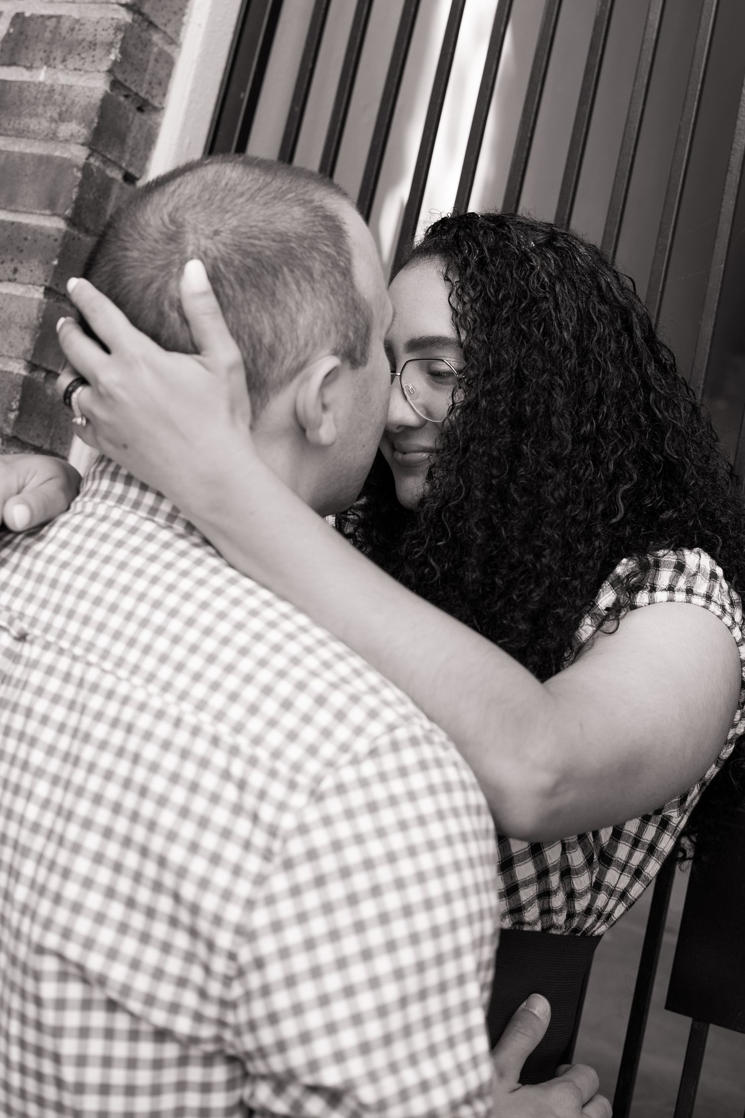 A couple shares an intimate moment kissing in black and white near a brick wall and metal fence. Taken by Row And Co Photography in FXBG VA