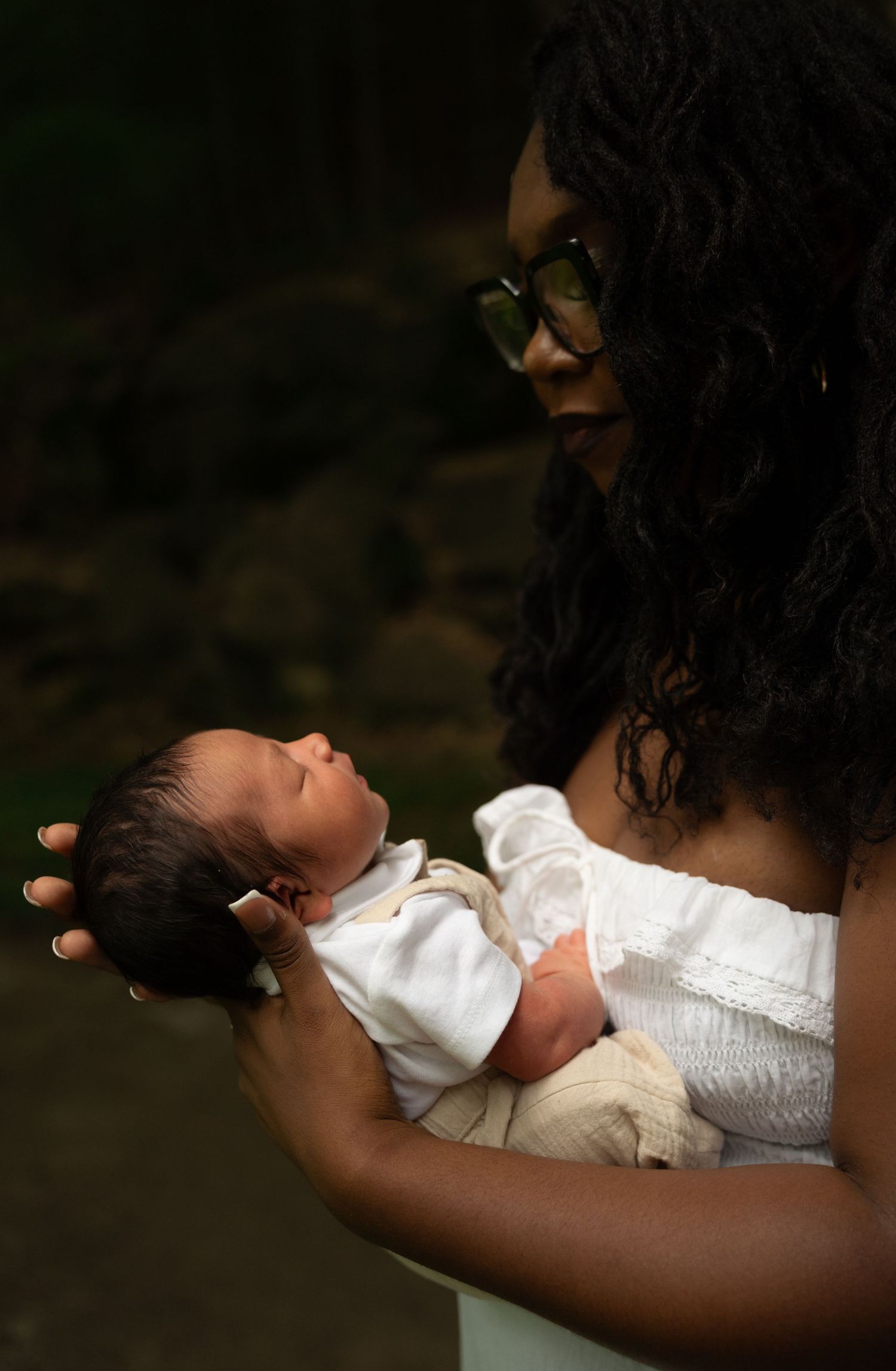 A Black woman with curly hair and glasses tenderly cradles her newborn baby dressed in white outdoors.