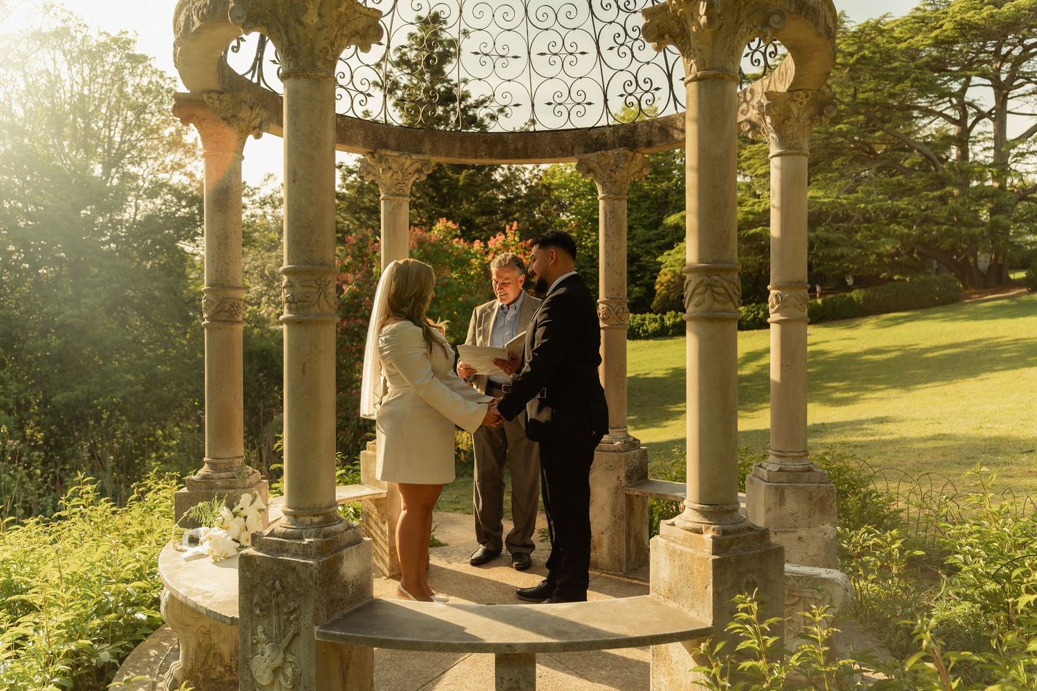 Couple exchanging vows under ornate stone gazebo in sunlit garden with officiant present.