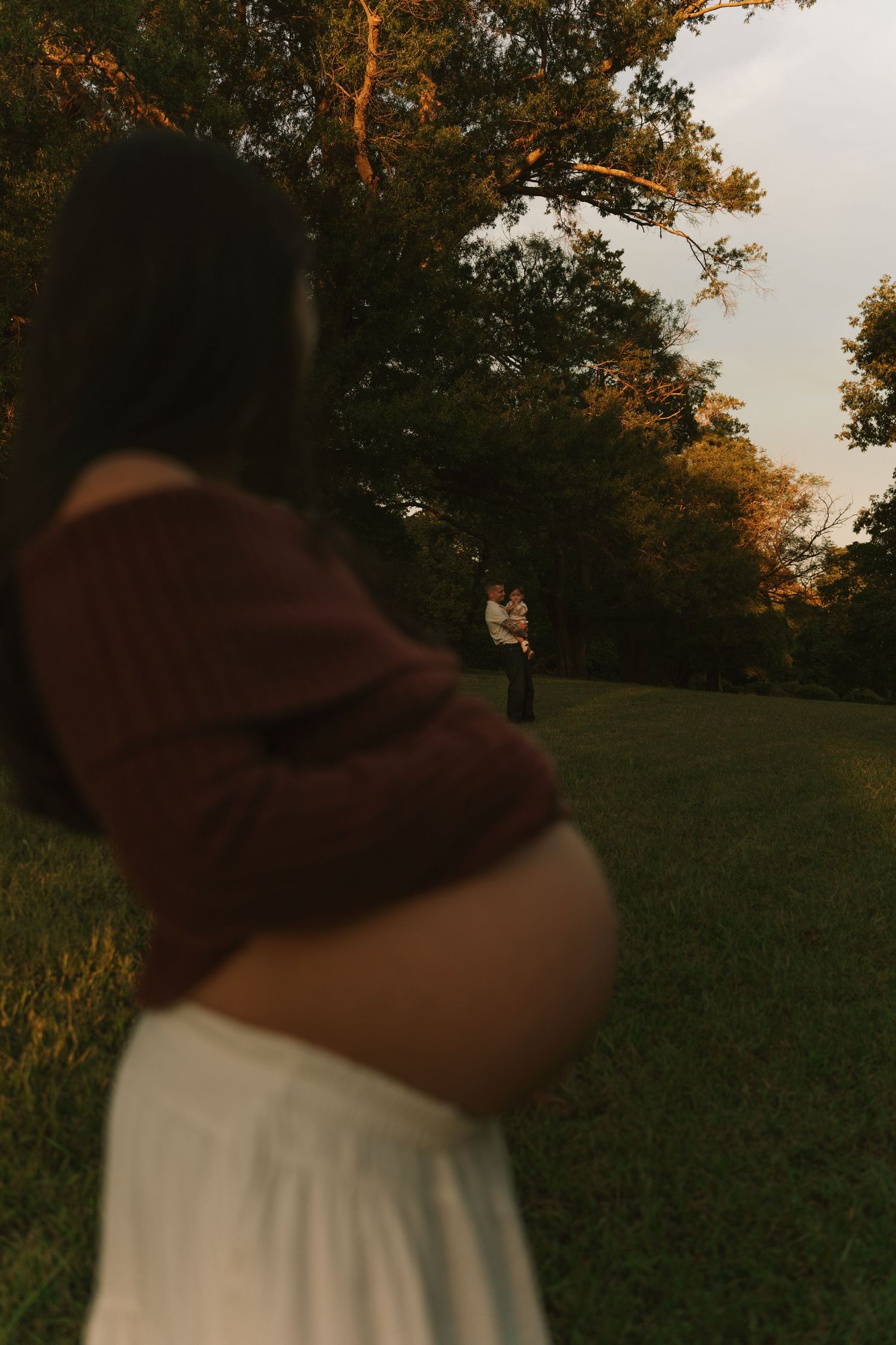 Blurred pregnant woman in burgundy top and white skirt stands in golden hour park with couple embracing in background.