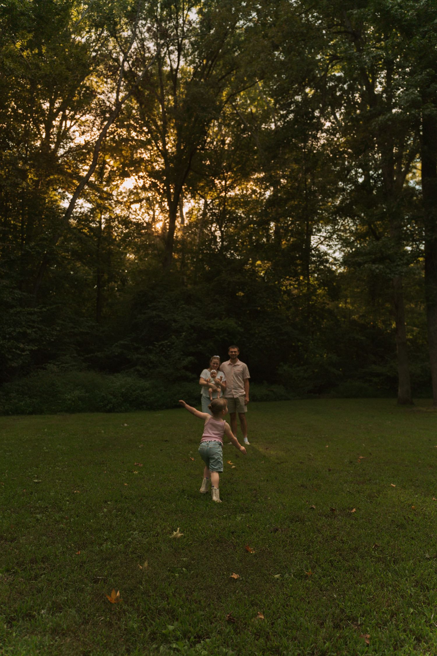A child runs toward two adults on a lush green lawn surrounded by trees at golden sunset hour.