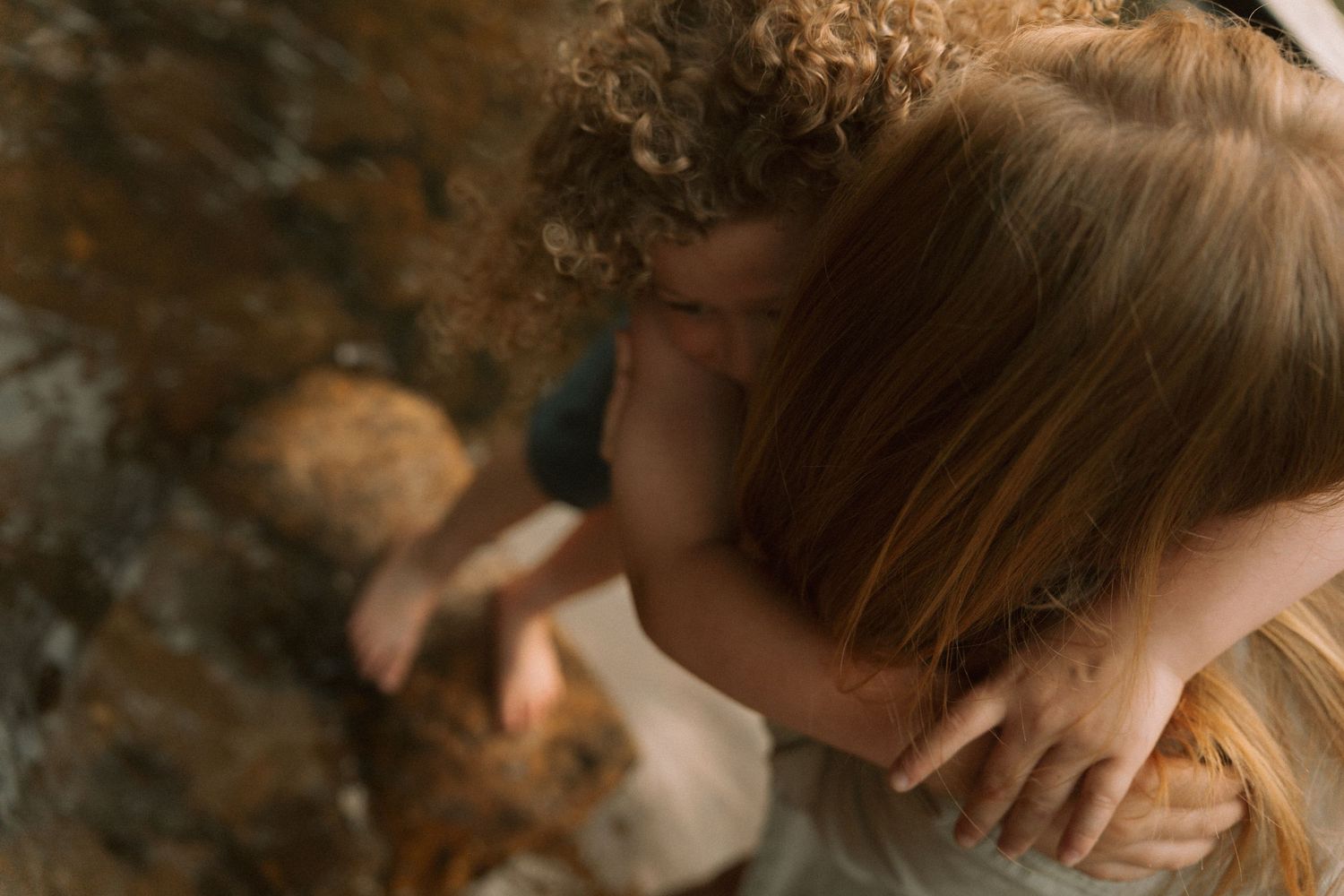 Two curly-haired children sharing a sweet embrace near a rocky surface, captured from above. Taken by Row And Co Photography in FXBG VA