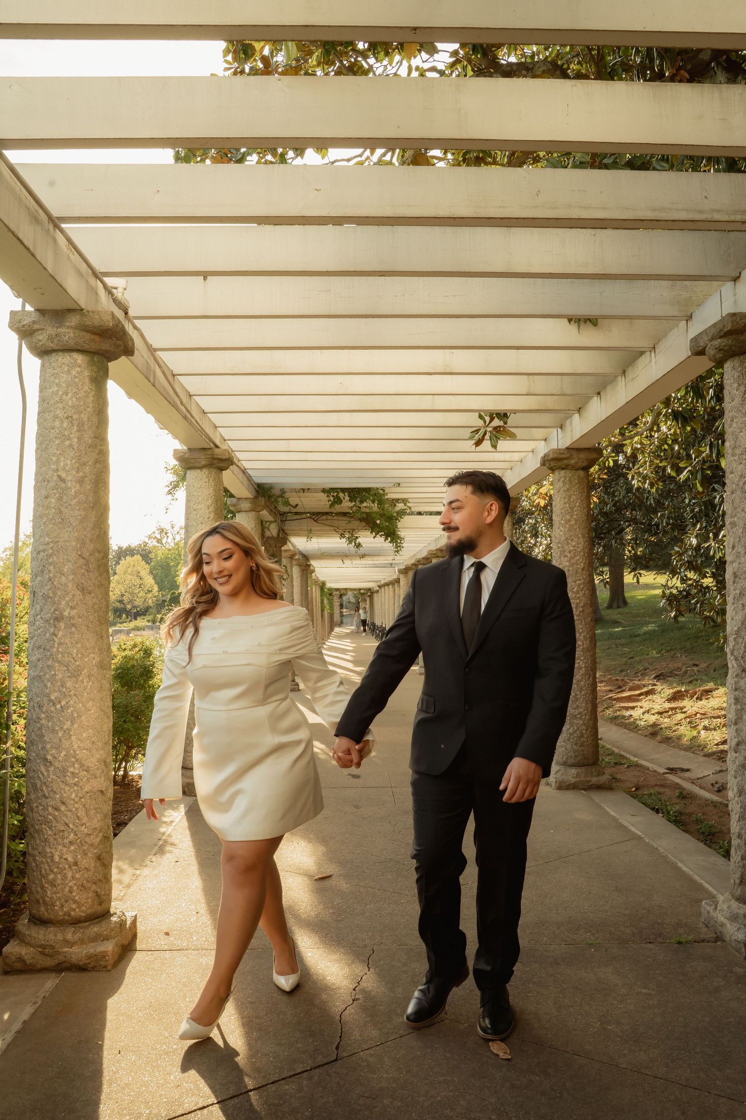 Couple in wedding attire holding hands and walking under a pergola, smiling joyfully in an outdoor garden setting.