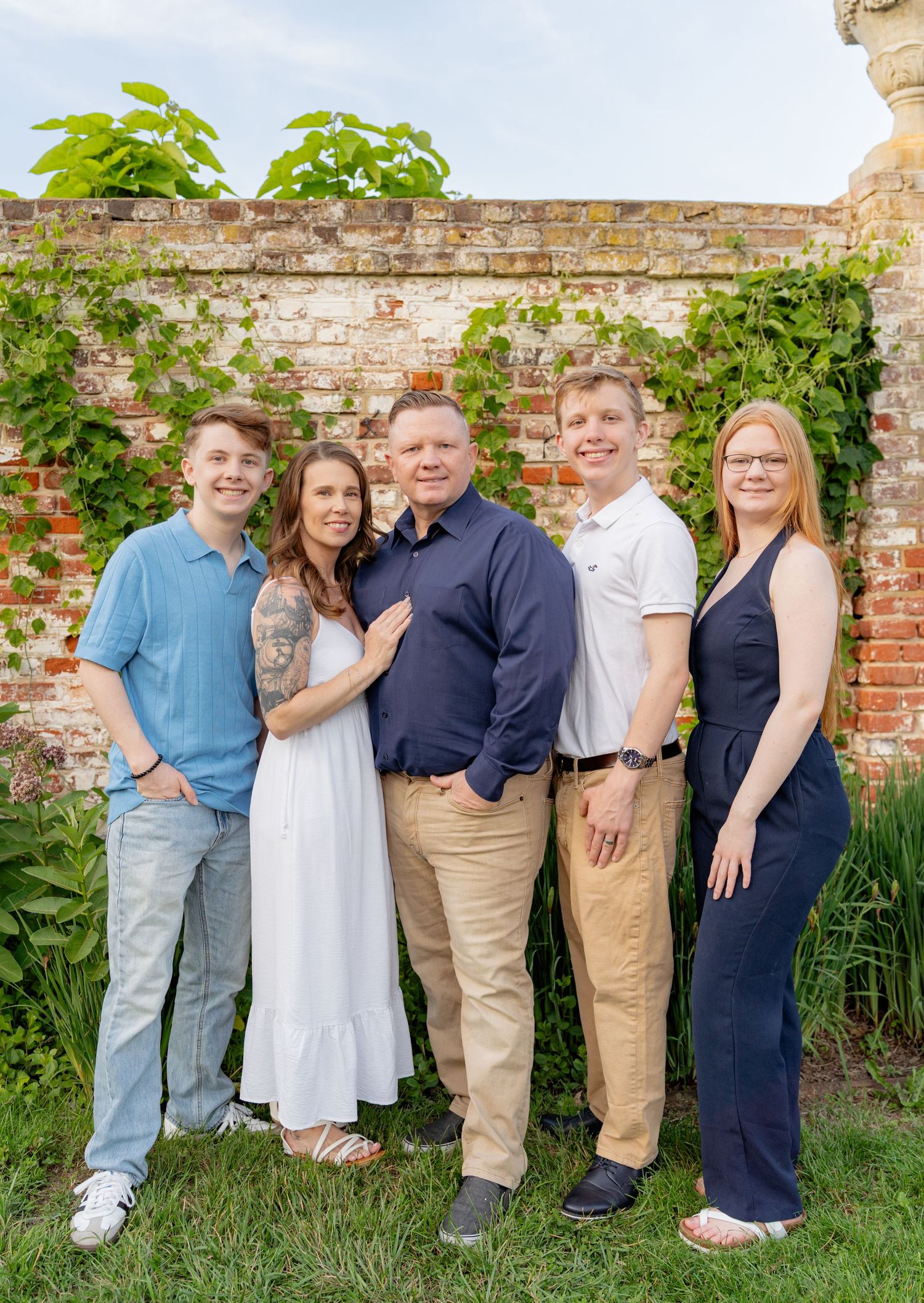A family of five poses together smiling in front of a rustic brick wall covered in green ivy. Taken by Row And Co Photography in FXBG VA