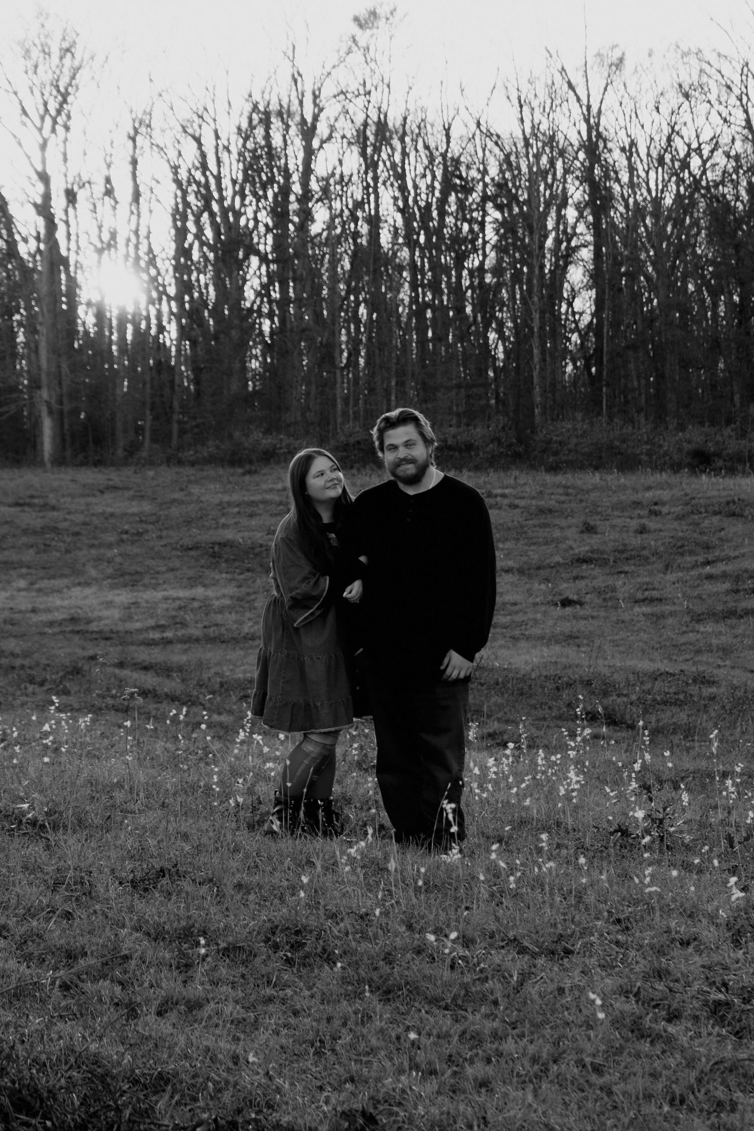 A couple stands together in a black and white photo in an open field at sunset.