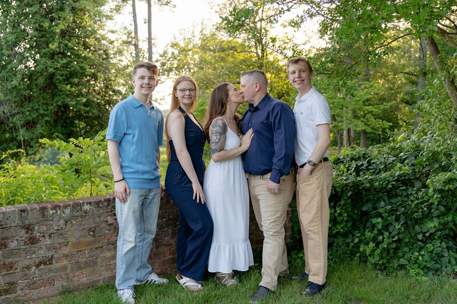 Family of five posing outdoors in a lush green garden, parents sharing a kiss while children stand alongside them. Taken by Row And Co Photo