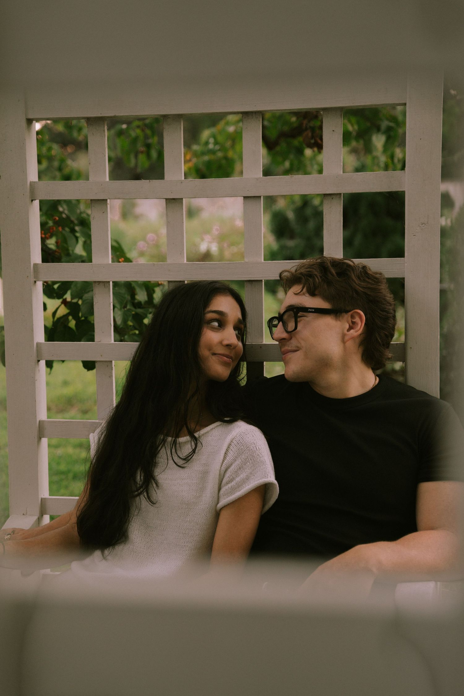 A young couple sitting together on a porch swing, smiling at each other in front of a white lattice backdrop. Row and Co Photography