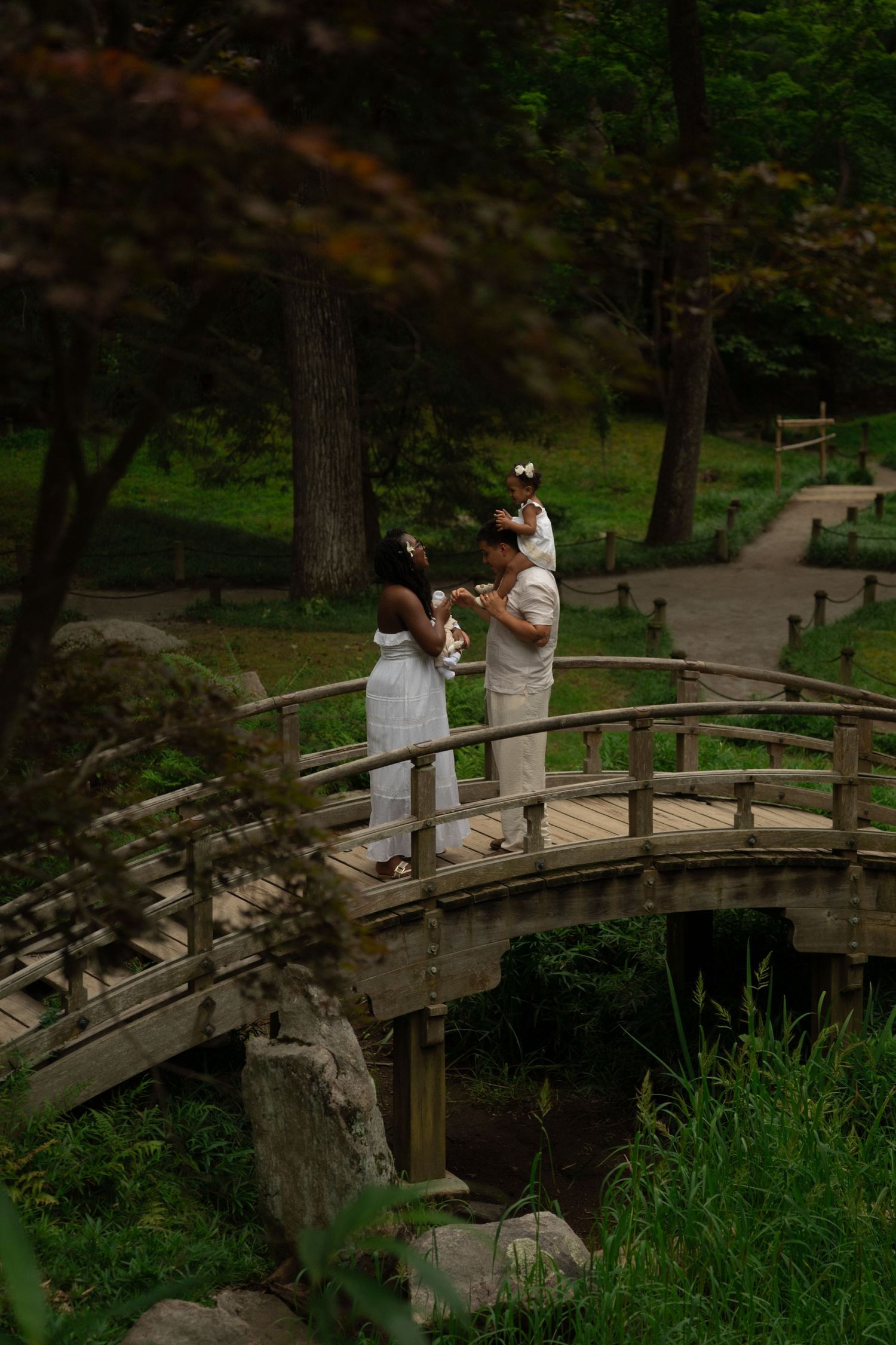 Family in white attire posing on a wooden arched bridge surrounded by lush green trees in a serene park setting.Row and Co Photography
