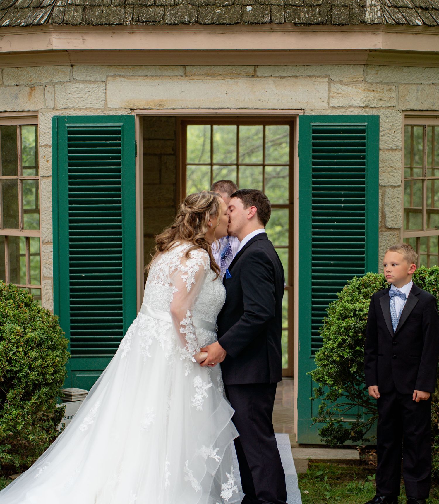 Bride and groom share first kiss at outdoor wedding ceremony beside green shuttered building with young boy watching. Row and Co Photography