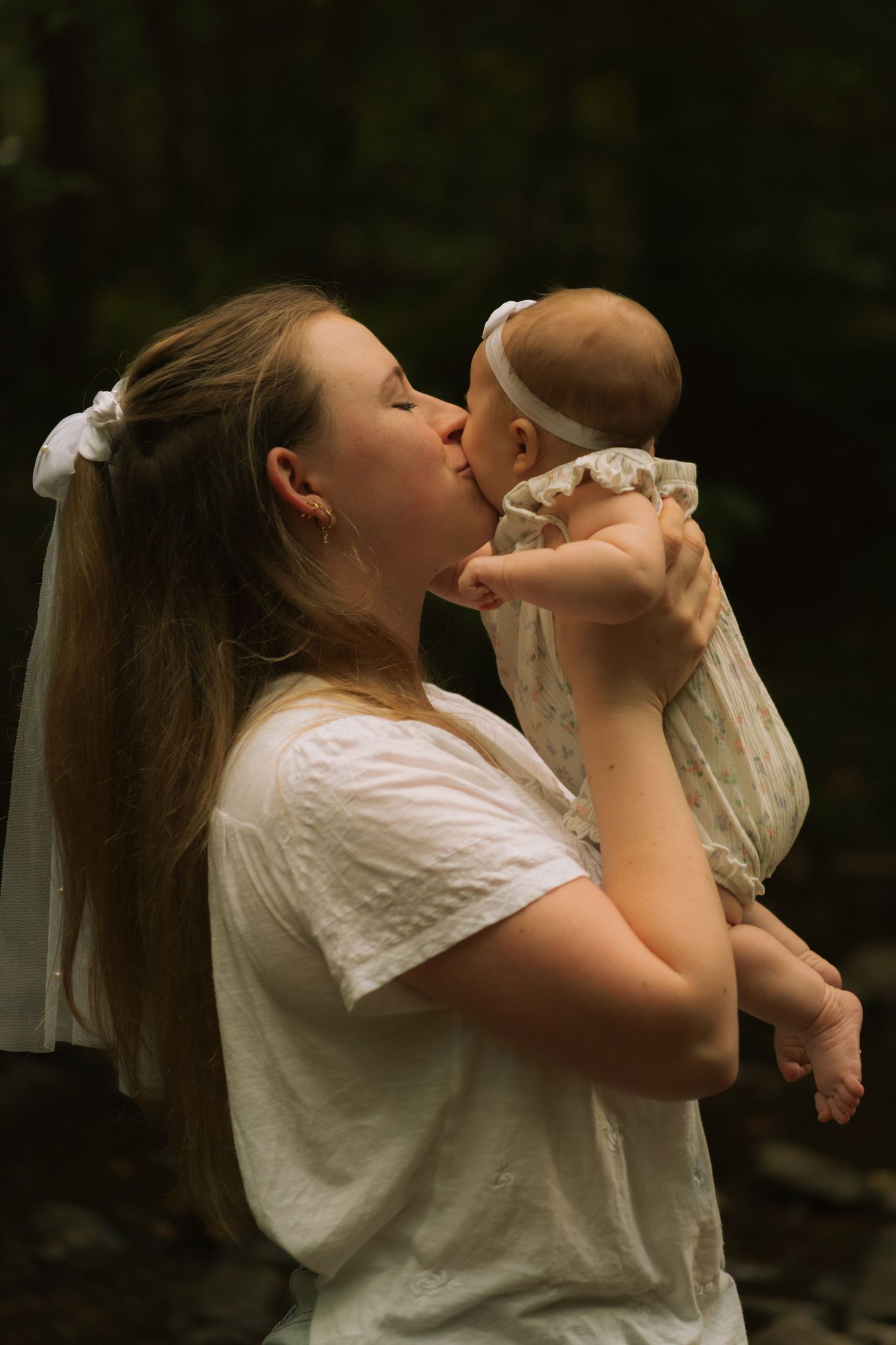 A young mother in a white dress kisses her newborn baby outdoors in a serene, dark green natural setting.