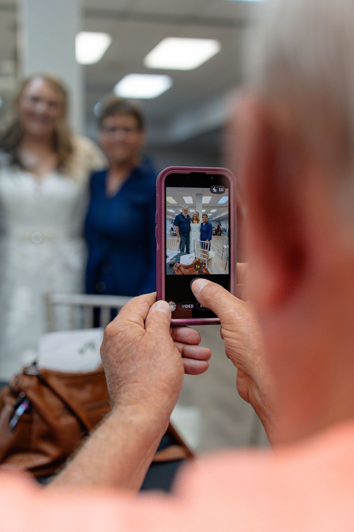 Person photographing two women with a pink-cased smartphone at an indoor event, capturing their portrait on screen.