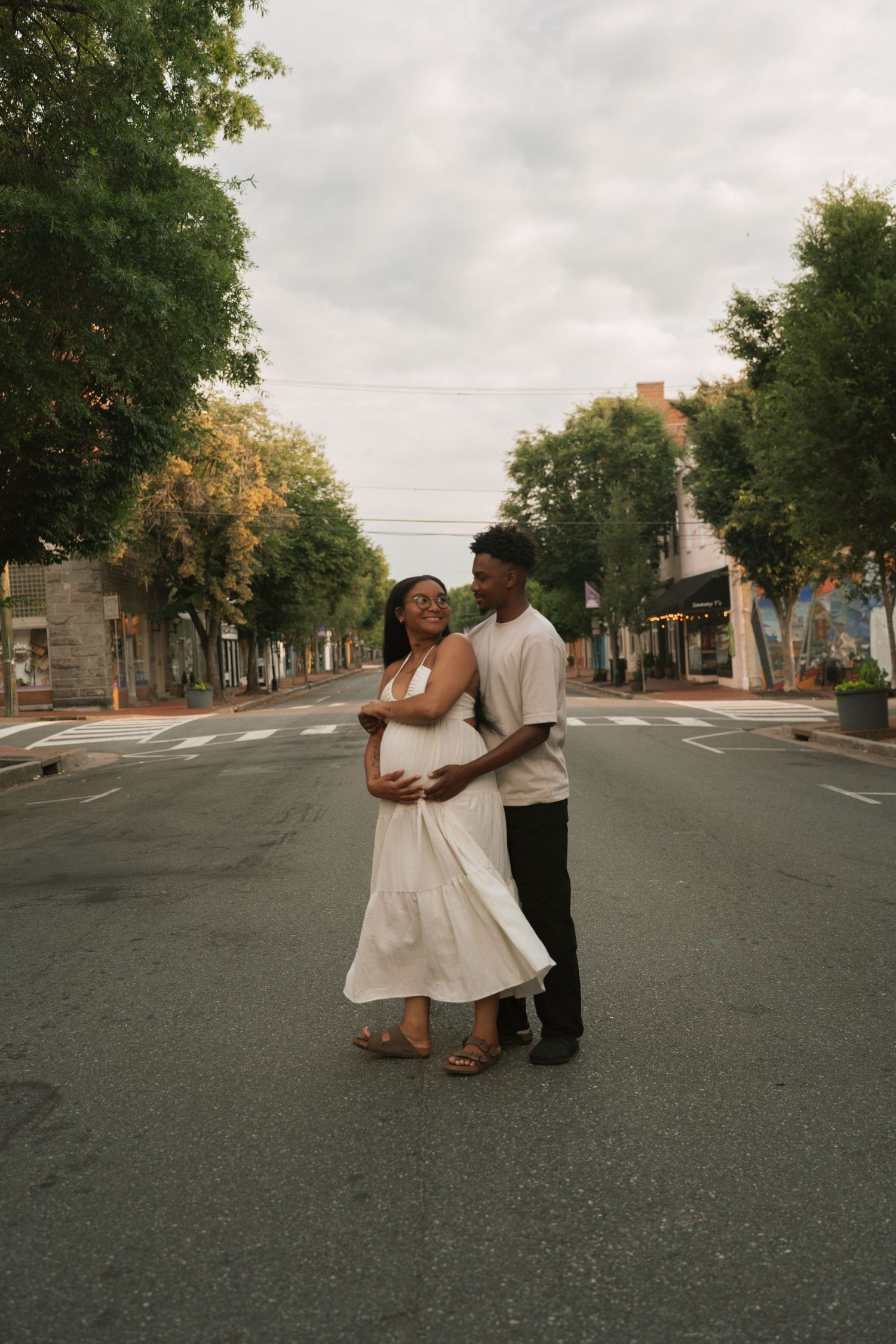 A couple embraces on an empty urban street lined with trees, the woman in a white dress cradling her baby bump.