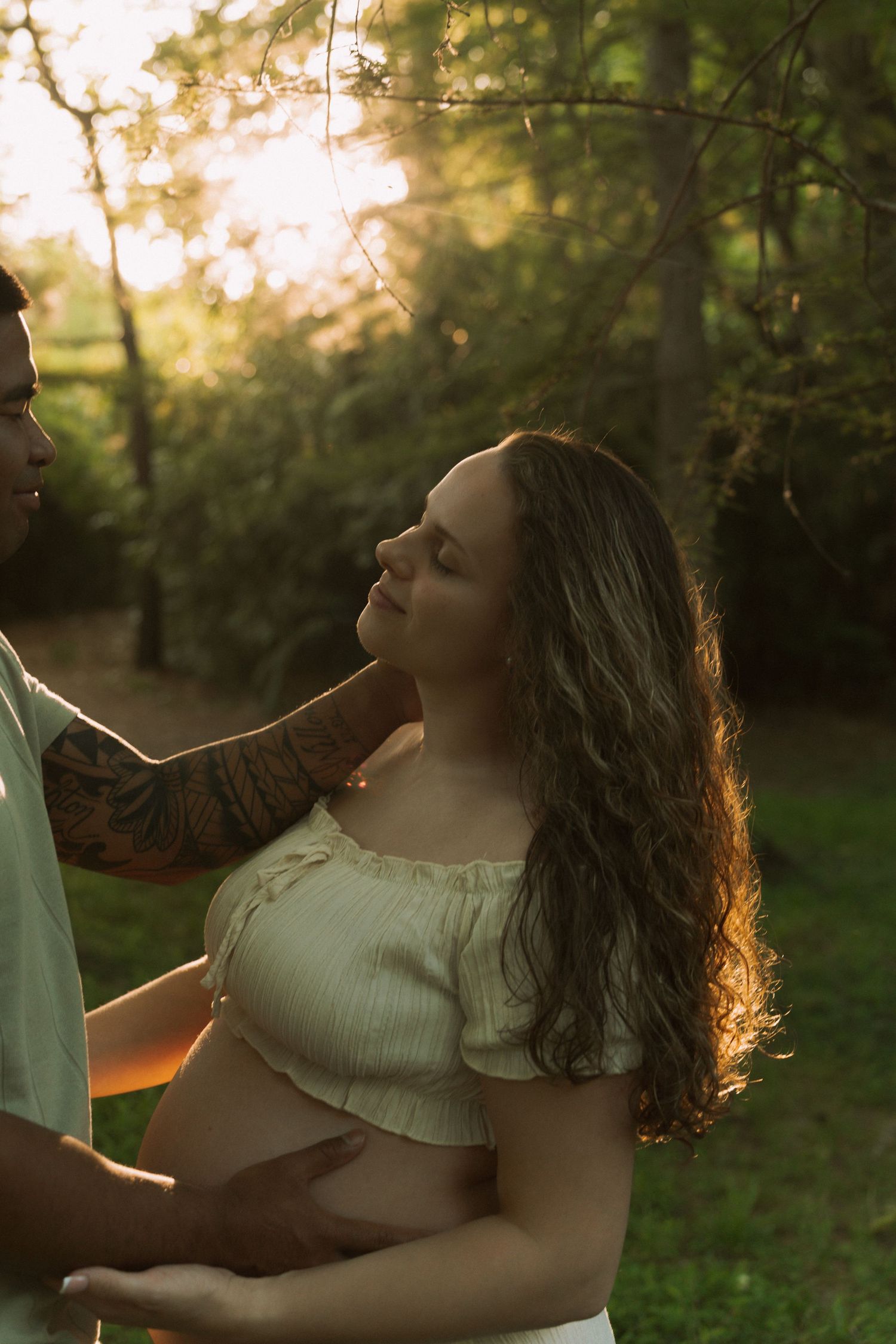 Pregnant woman with long hair stands outdoors at golden hour as partner with tattooed arm gently touches her face.