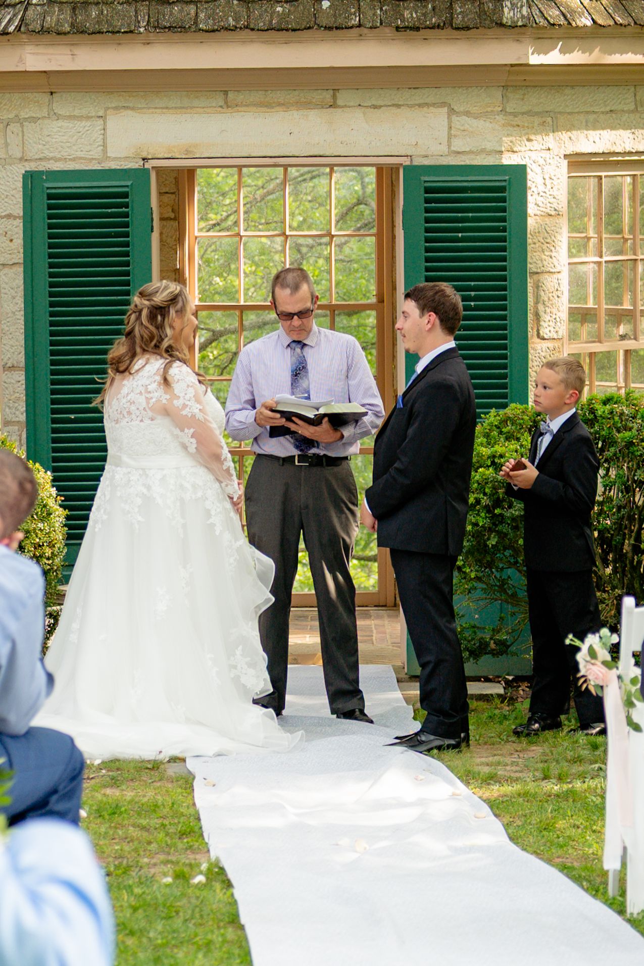 Outdoor wedding ceremony with bride in white gown, groom in suit, and officiant reading vows by stone building. Row and Co Photography