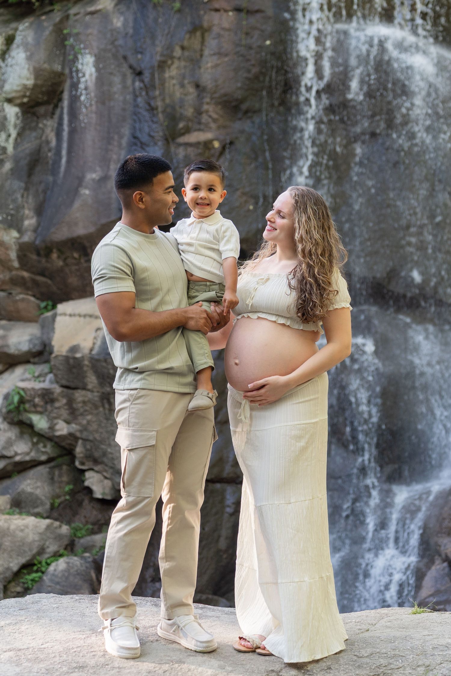 Expecting couple with toddler son posing in front of a waterfall during a maternity photo session. Taken by Row And Co at Maymont RVA