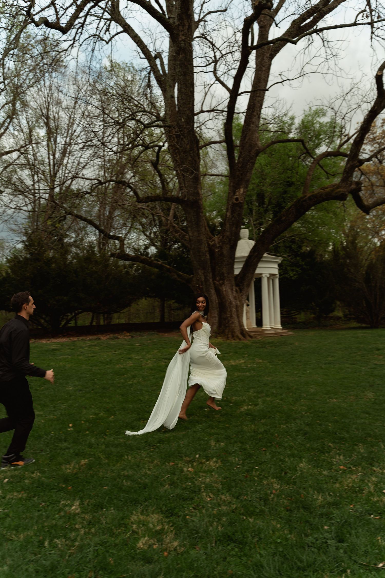 Bride in white gown runs toward groom on lush green lawn with large tree and white gazebo backdrop. Taken by Row And Co Photography in FXBG