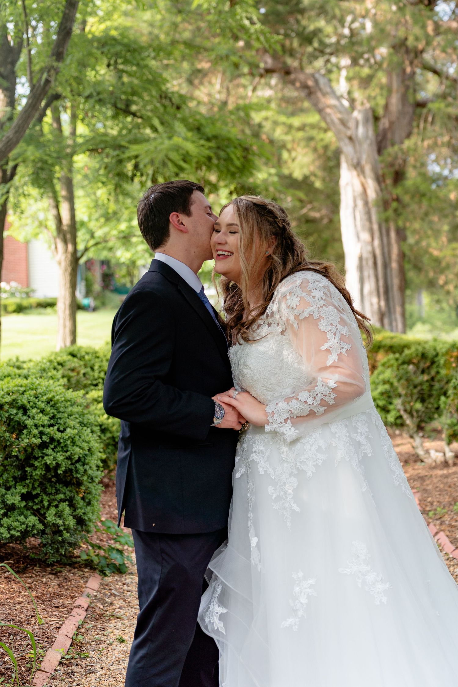 A bride in lace gown smiles as groom kisses her cheek in a garden setting.