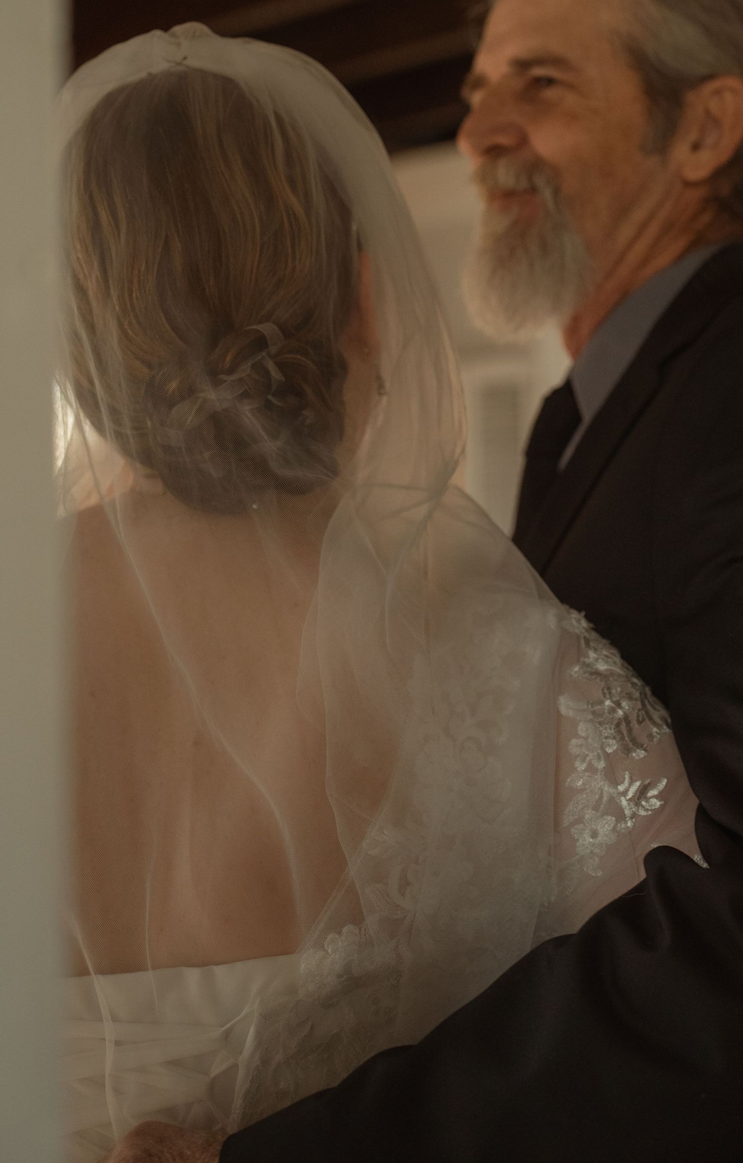 Bride in white lace wedding dress and veil stands with bearded man in dark suit, sharing a tender moment.